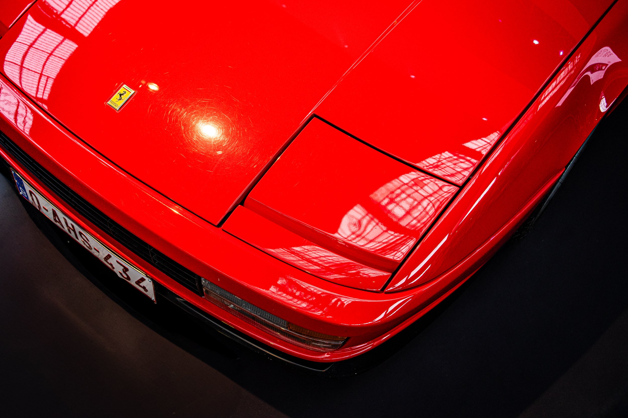 Close-up of the front of a red Ferrari sports car, showing the hood with a Ferrari badge and the front license plate, with reflections of a large glass ceiling.
