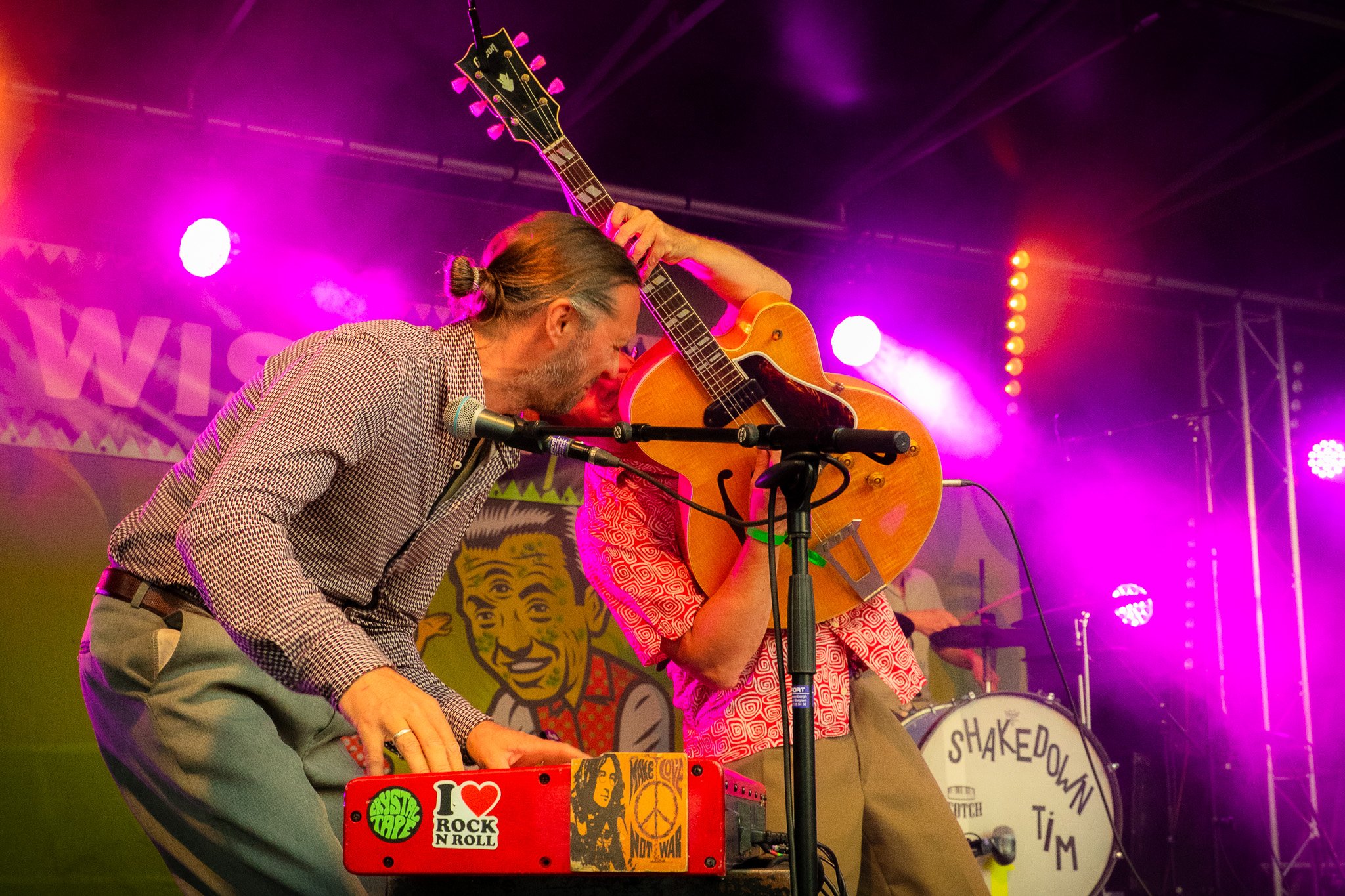 A man and a woman playing musical instruments on stage with colorful lights. The man is playing a keyboard, and the woman is playing a guitar. The stage has vibrant purple and pink lighting, and there are stickers on the keyboard including peace sign