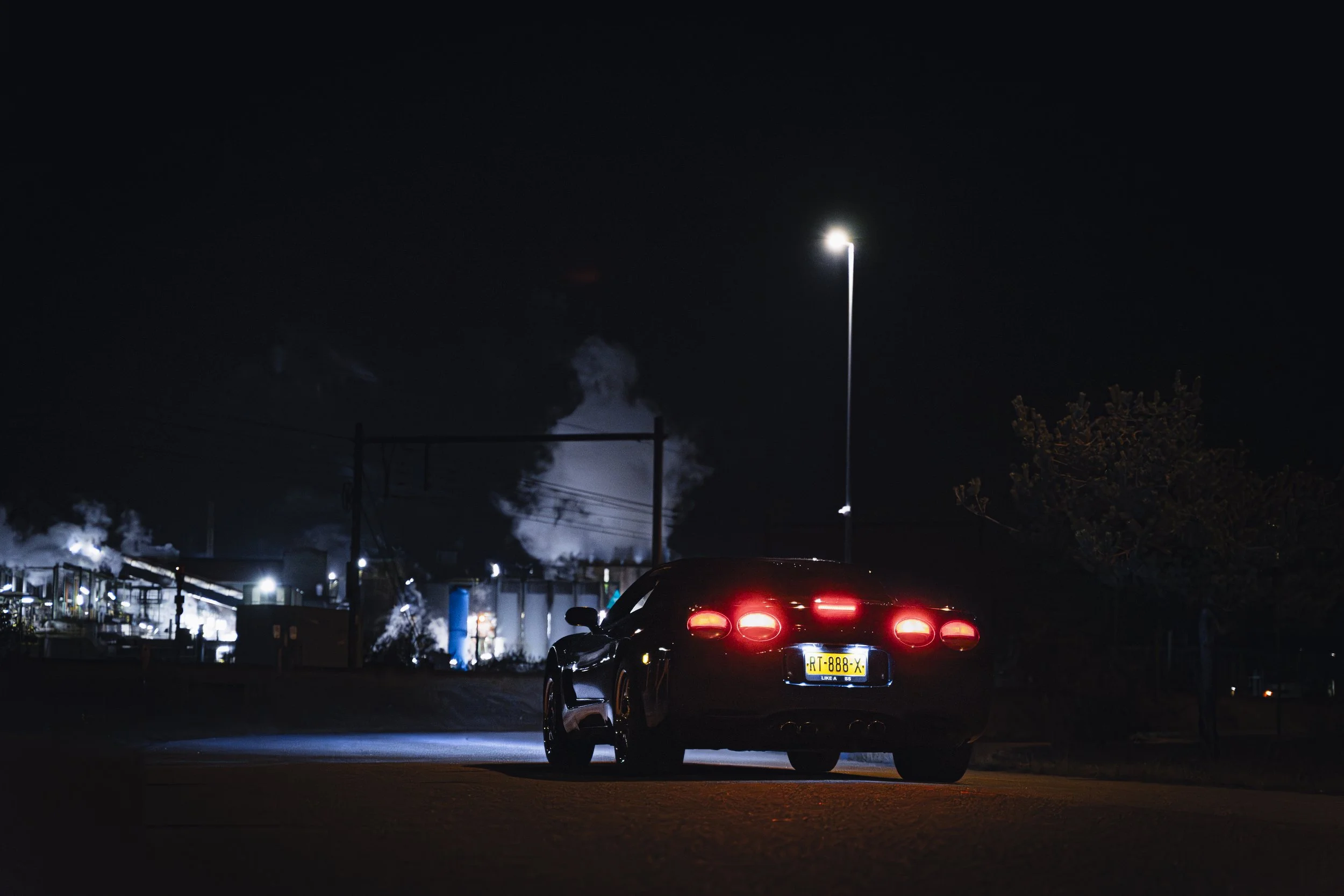 A black sports car with its rear lights on is parked under a streetlight at night. In the background, an industrial area with smoke rising from buildings is visible.