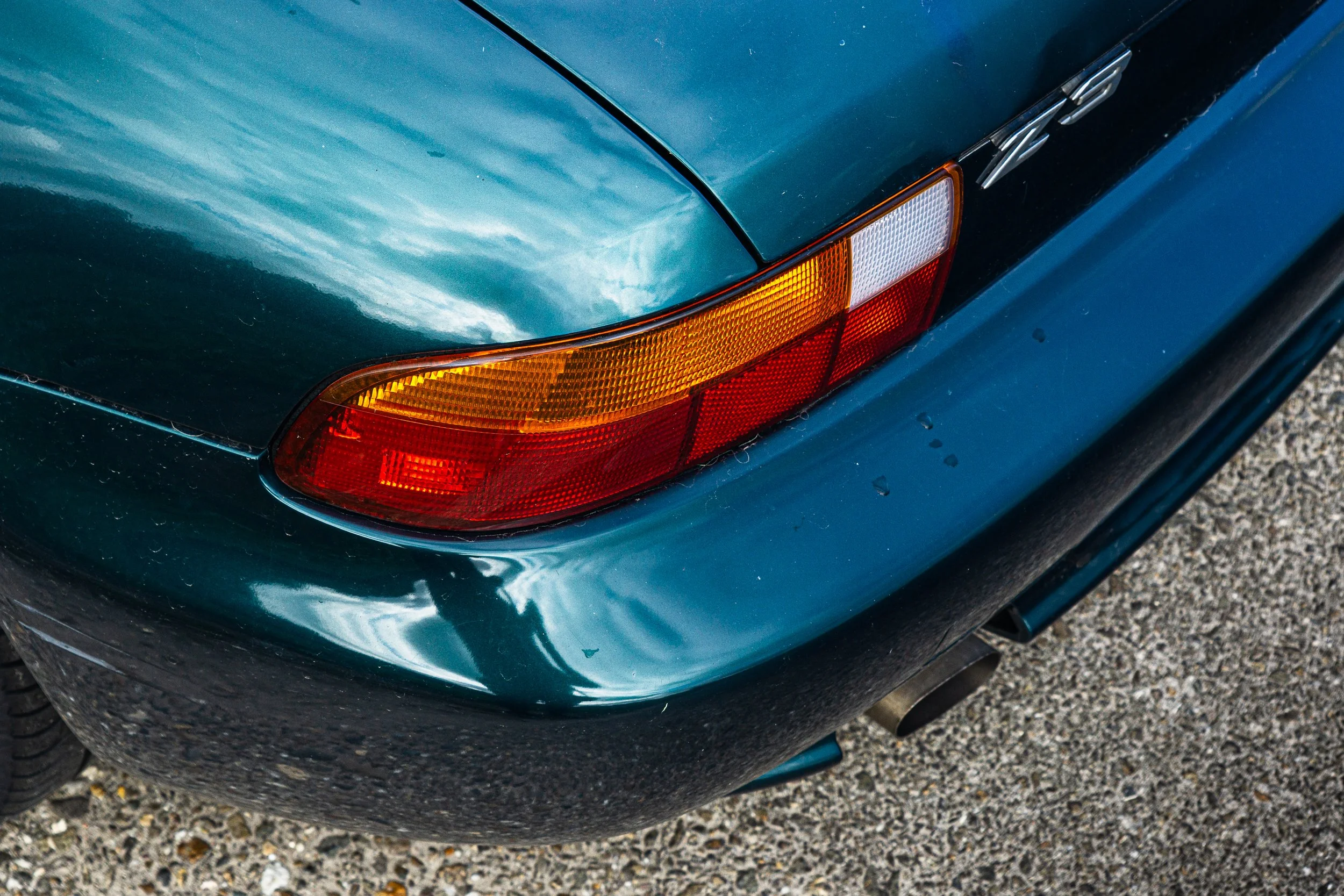 Close-up of the rear right corner of a teal classic car showing the tail light, bumper, and exhaust pipe.