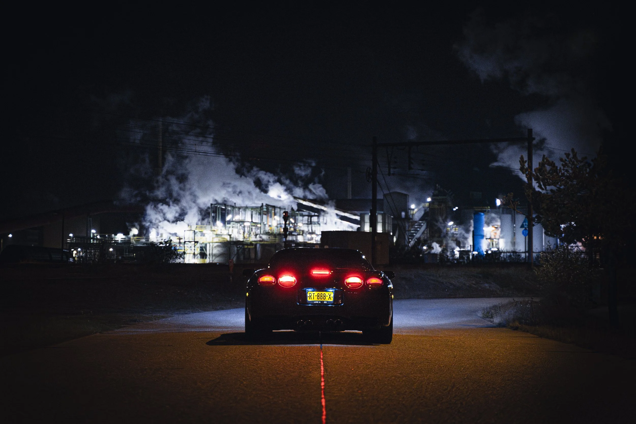A black sports car with red taillights parked on a road at night, with an industrial plant emitting smoke in the background.