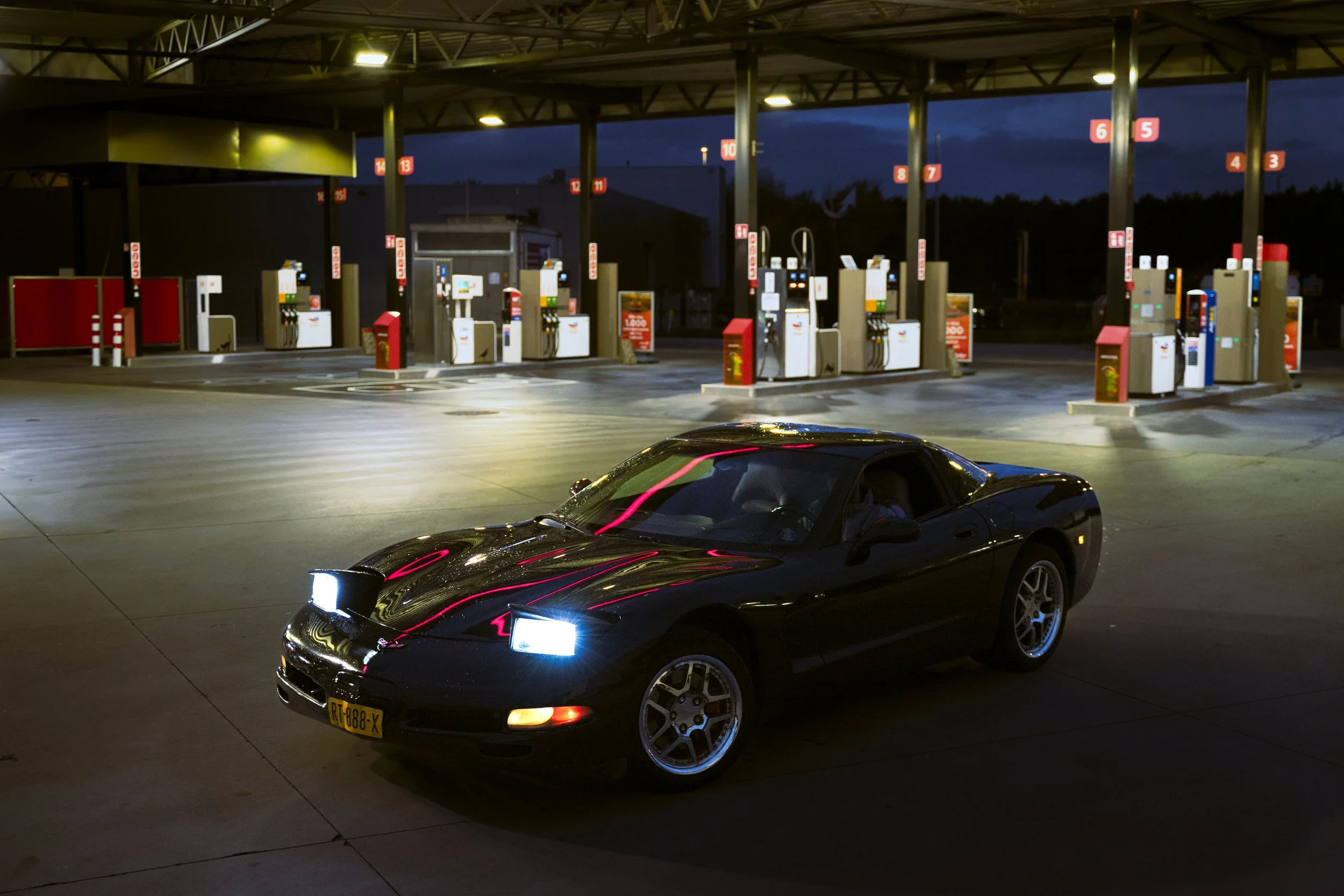 Black sports car parked at a gas station at night with illuminated pumps and a dark sky in the background.