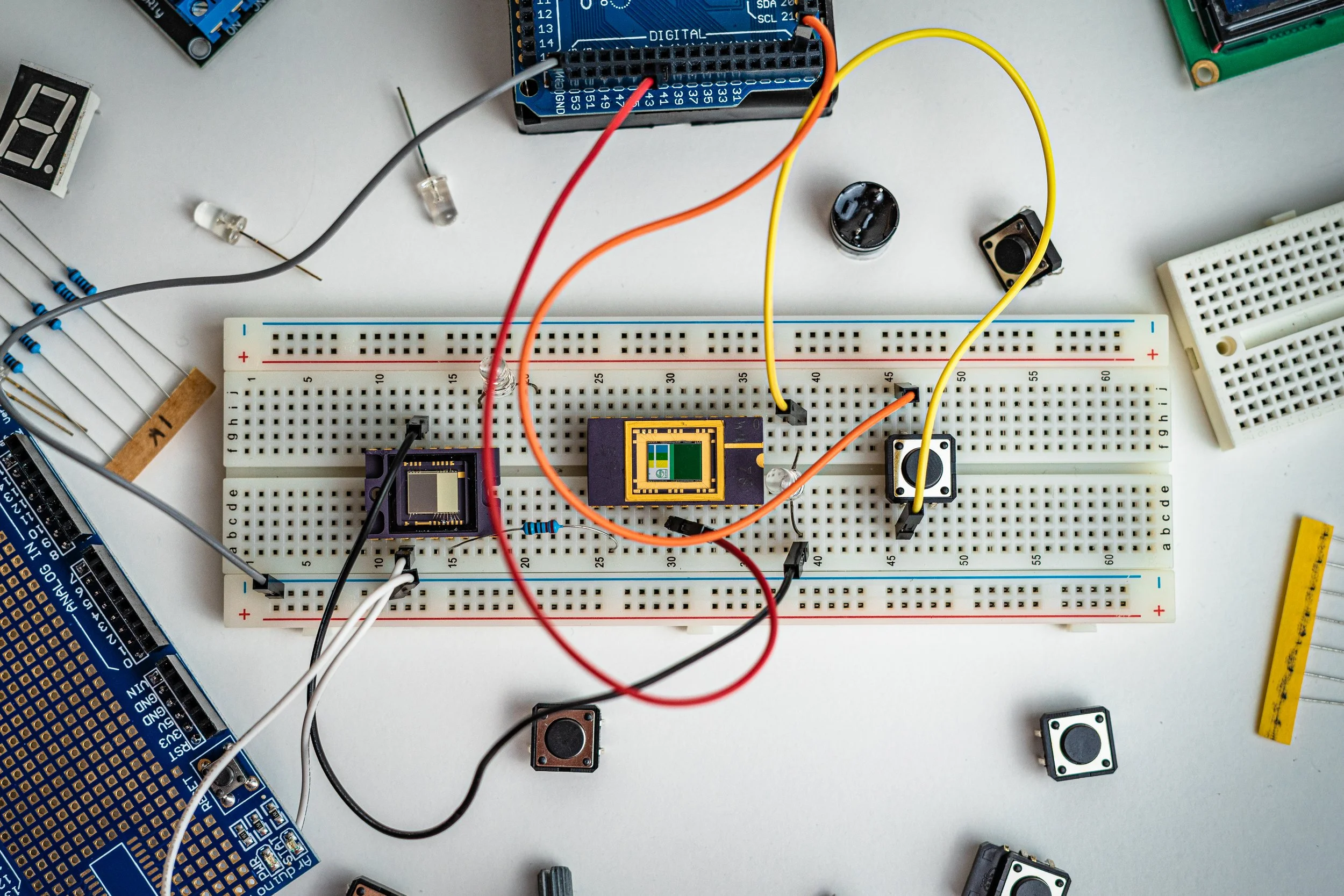 A breadboard with various electronic components and wires, including microprocessors, resistors, switches, and a digital display, on a white surface.
