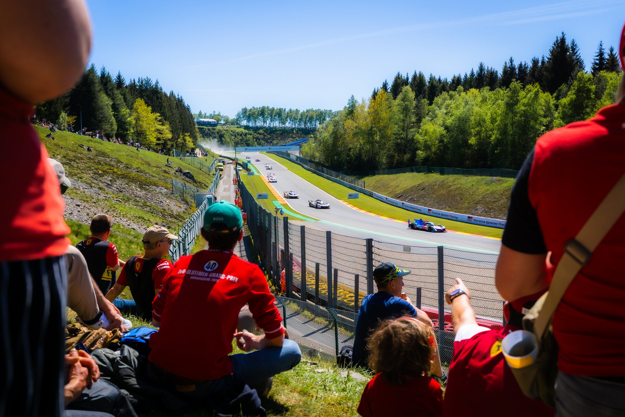 Spectators watching a car race from the grassy hillside at a motorsport circuit on a sunny day, with racing cars speeding down the track.