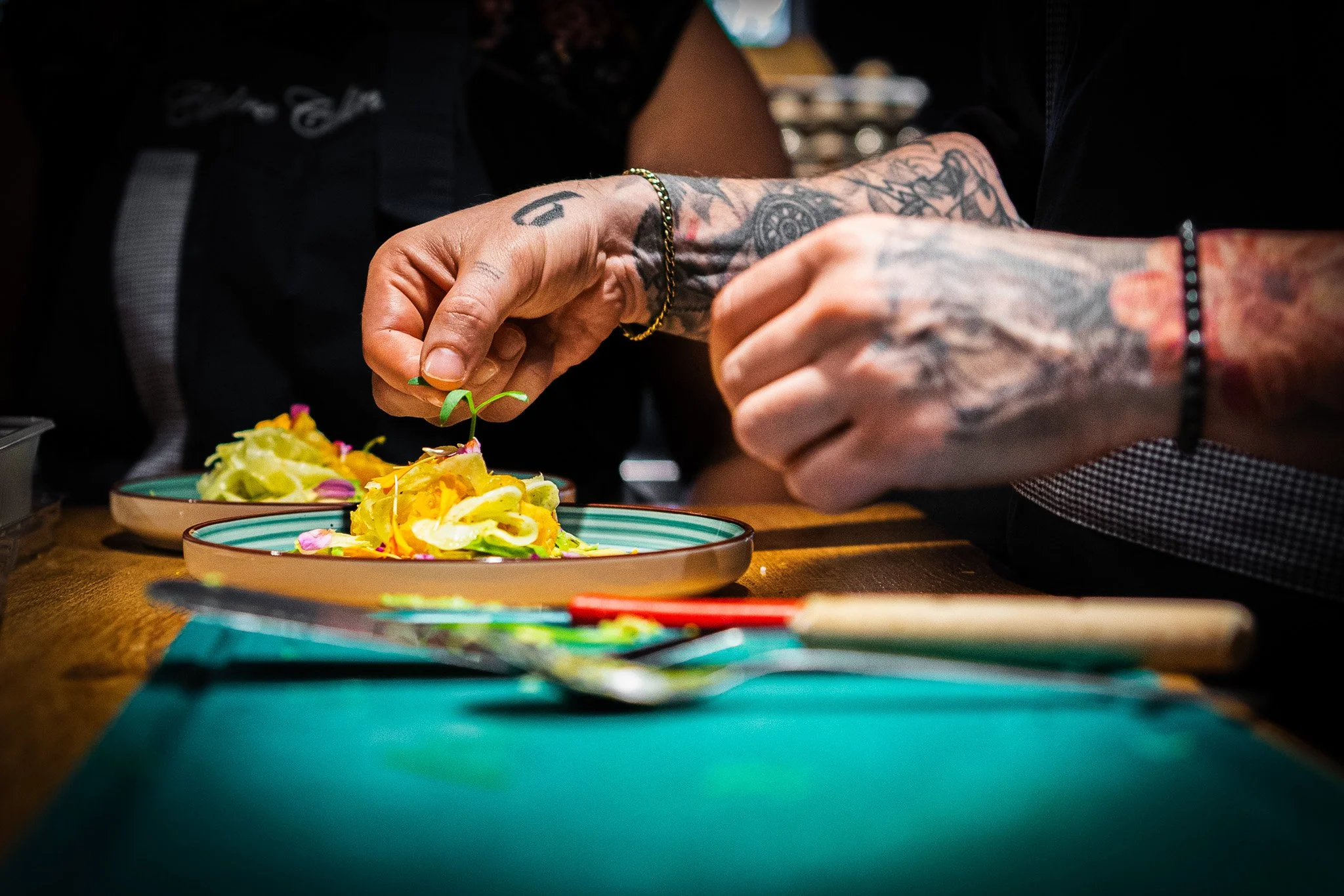 Close-up of a person with tattooed arms preparing a colorful dish with yellow, pink, and green ingredients on a plate, on a wooden table.
