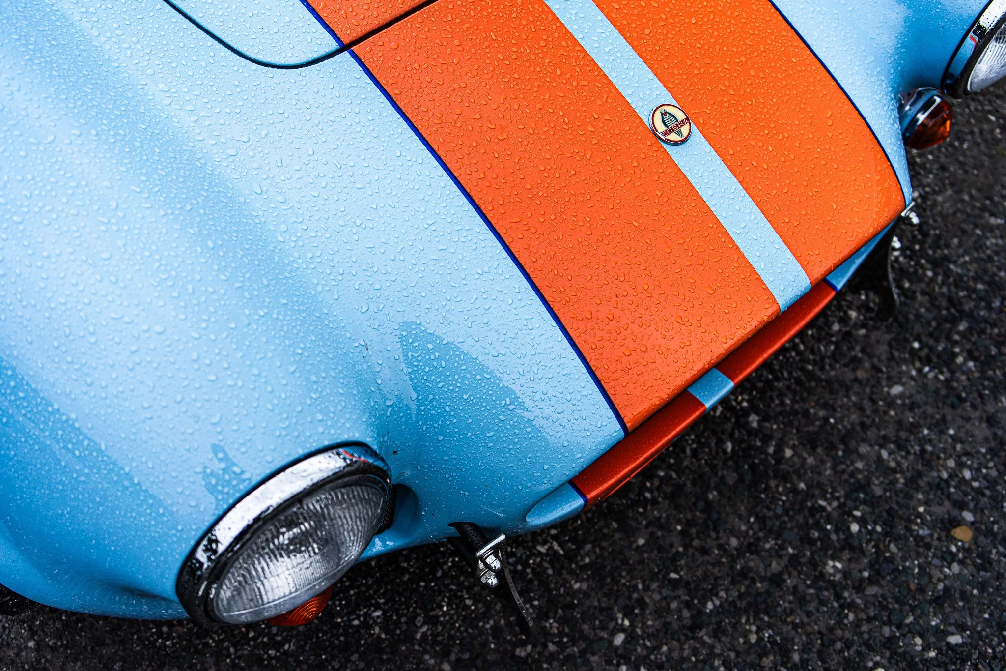 Close-up of a vintage light blue and orange racing car's front end with water droplets on the surface, a round headlight, and a small side mirror.