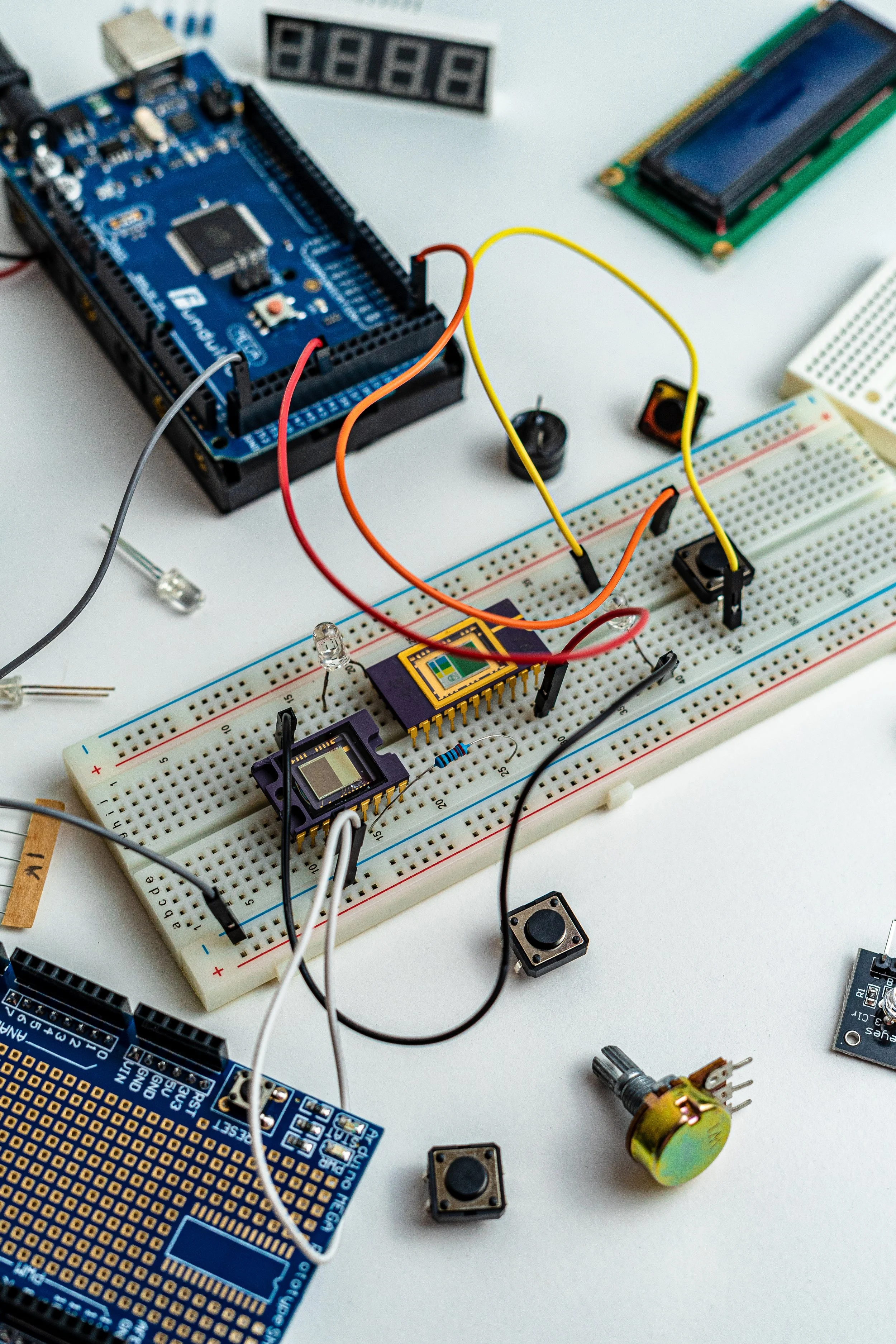 An electronics project setup with an Arduino microcontroller, a breadboard with various electronic components and wires, an LCD display, a couple of push buttons, a 7-segment display, a relay, and other electronic modules on a white surface.
