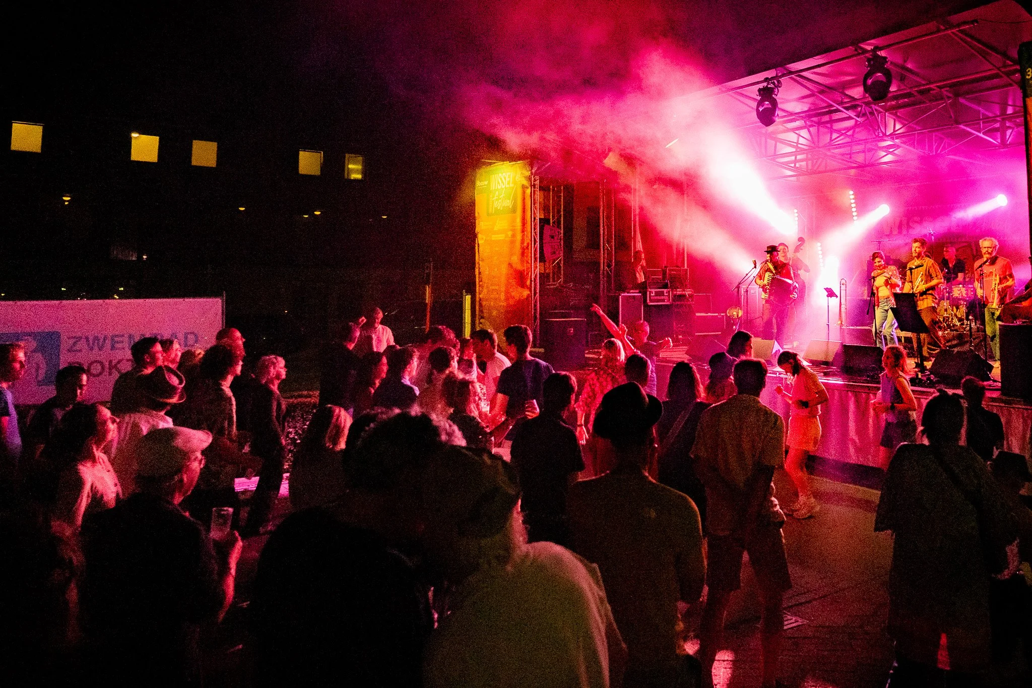 A stage with musicians performing at night, pink and purple lighting, and an audience watching in front.