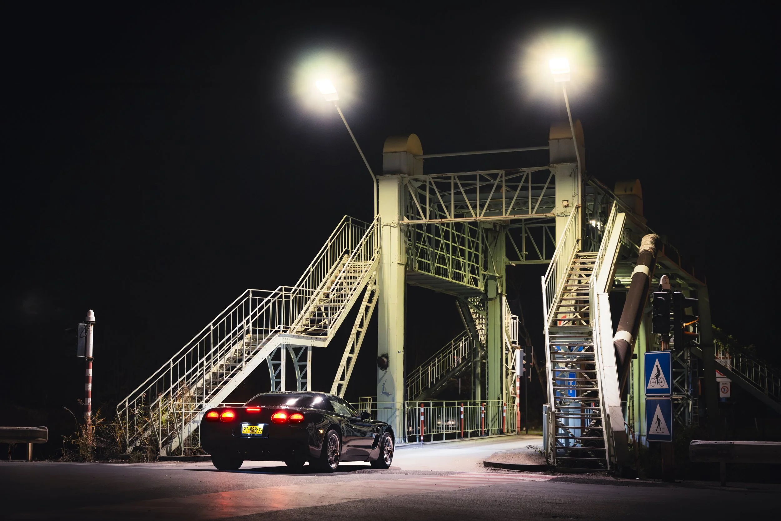 A black sports car parked beneath a metal pedestrian and bicycle bridge at night, illuminated by two bright streetlights.