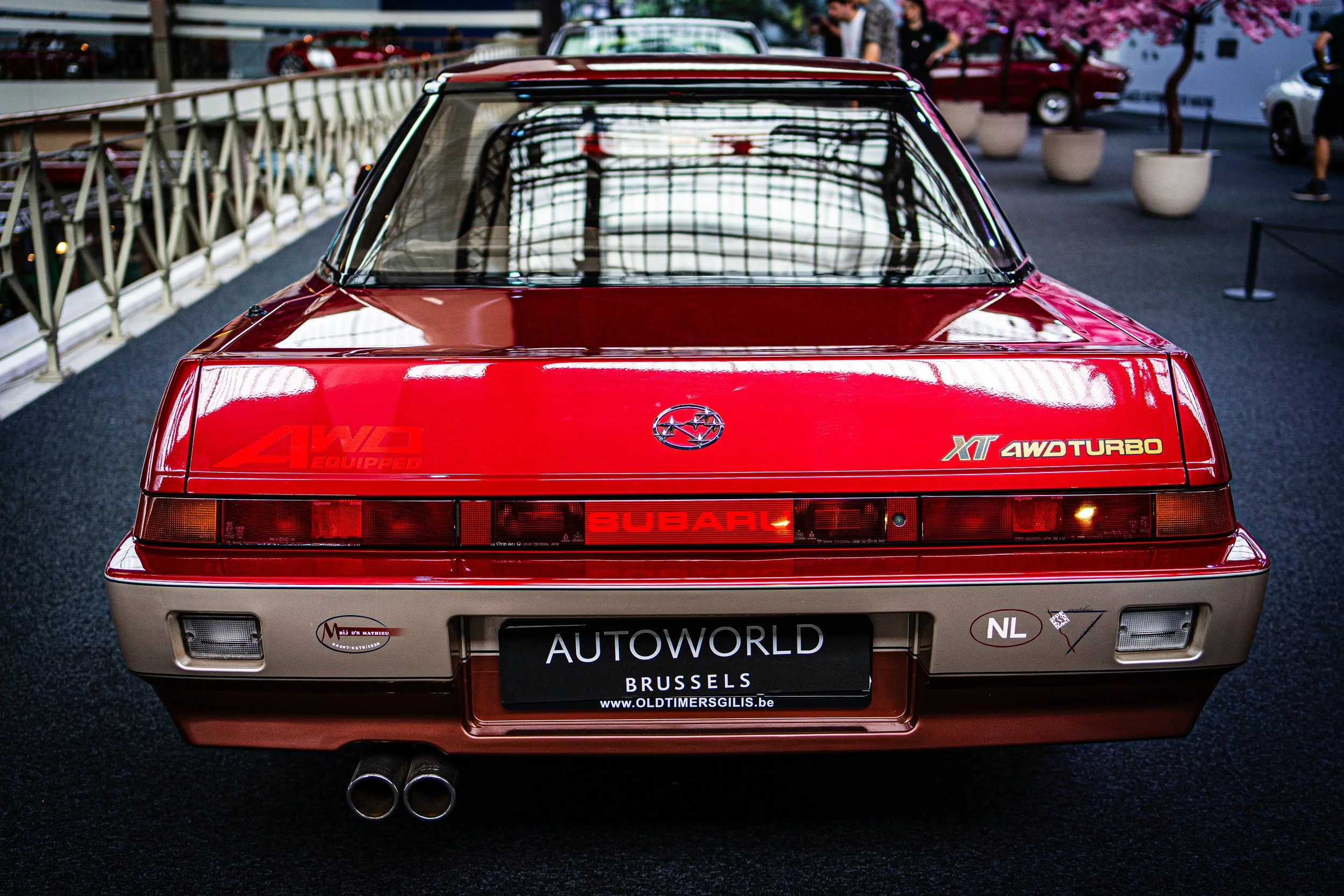 Red vintage Subaru XT car with dual exhaust pipes on display at an auto show, with black carpeted floor and potted plants in the background.