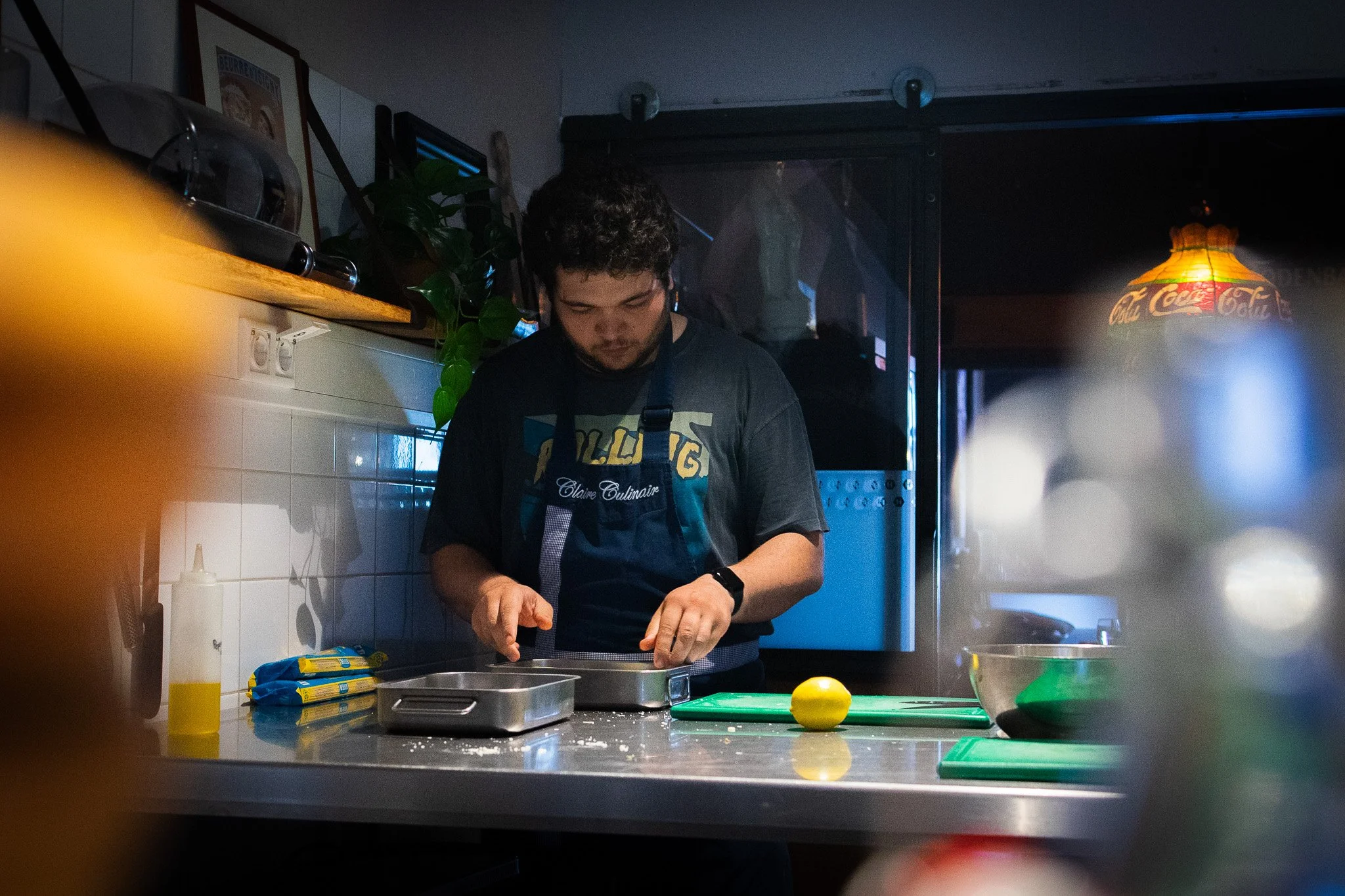 A young man with curly dark hair working in a kitchen, wearing a black apron, standing over a counter with a lemon and food items, amidst kitchen appliances and utensils, illuminated by dim lighting and a decorative hanging lamp.