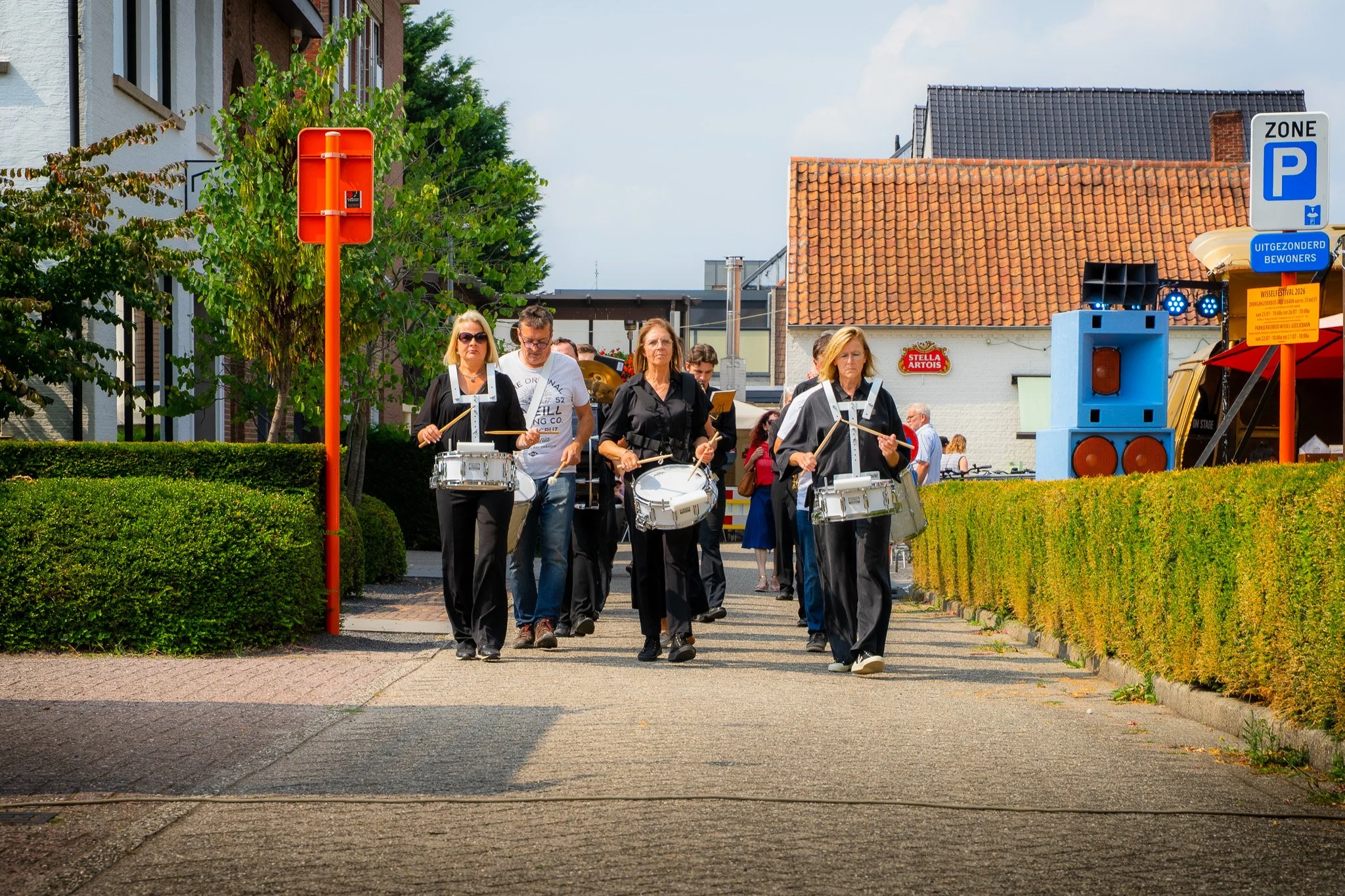 Group of musicians marching in an outdoor event, playing drums and percussion instruments, with green bushes, trees, and buildings in the background.