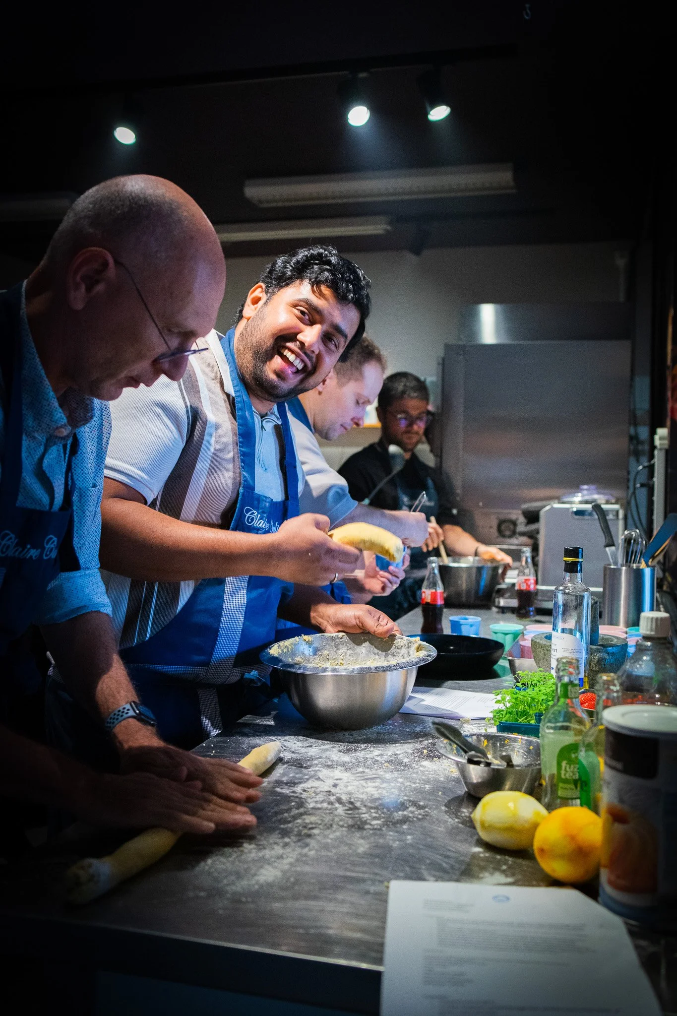 Group of people in a cooking class or kitchen, preparing food at a counter with various ingredients and utensils, smiling and working together.