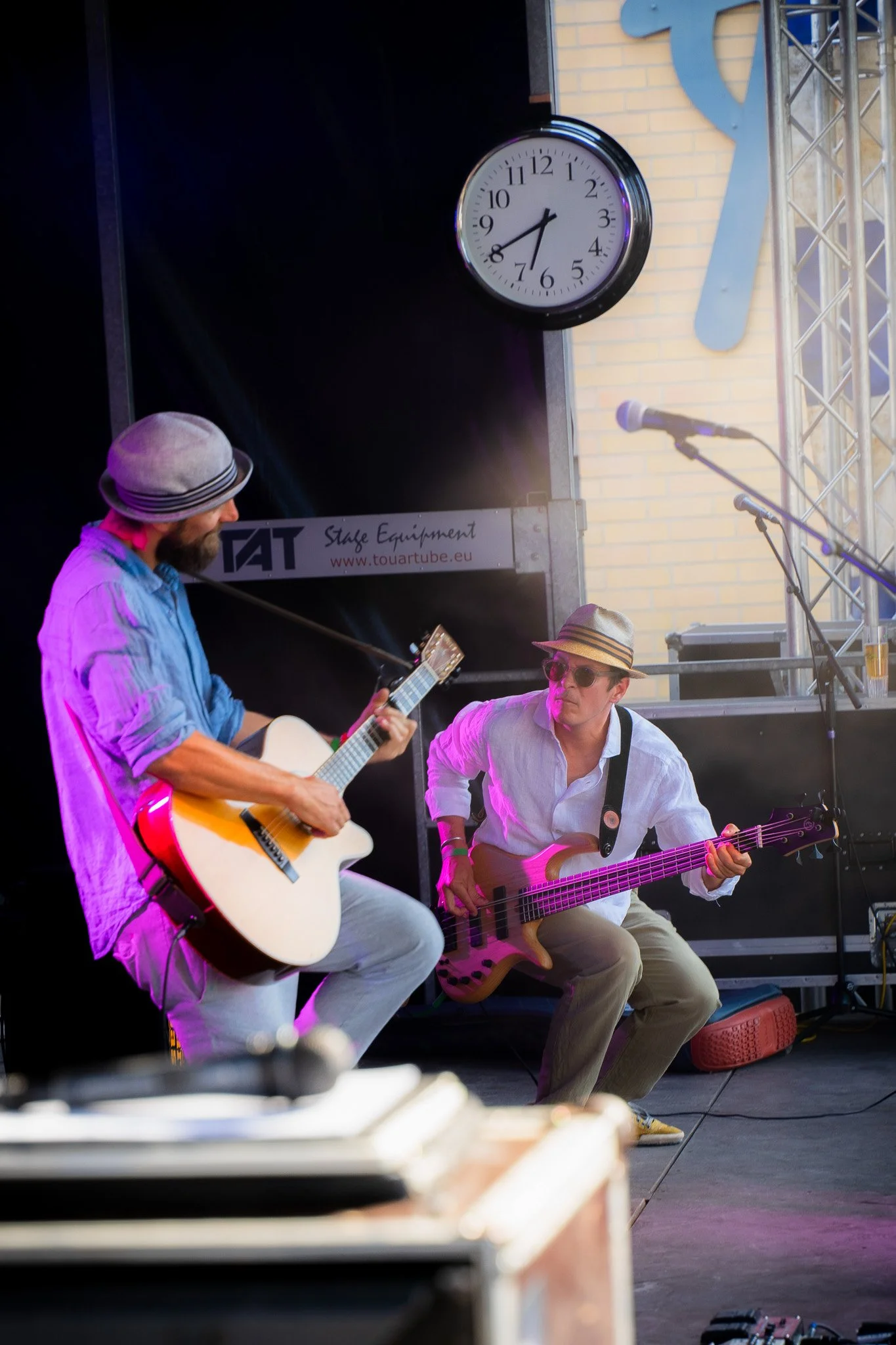 Two musicians playing guitars on an outdoor stage with a large clock and a microphone stand in the background.