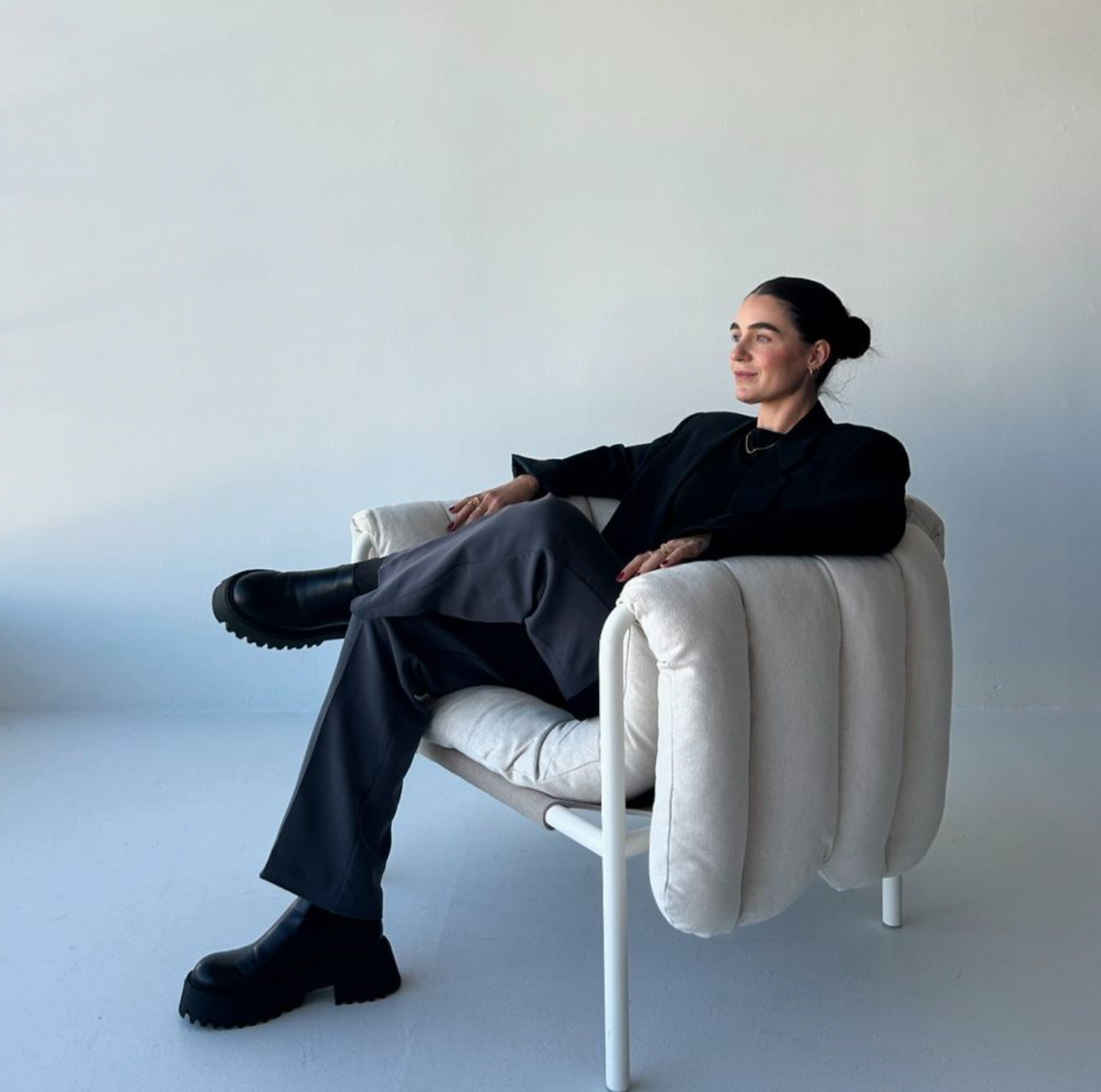Jemma Gwilliam, Woman sitting in a cream armchair, wearing black boots, gray pants, and a black blazer, with her hair in a bun, against a plain white wall.