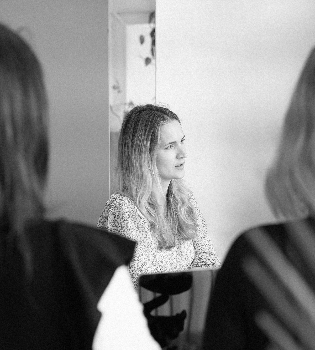 A woman with long wavy hair and a patterned blouse sitting at a table, reflected in a mirror, with two other women partially visible in the foreground.