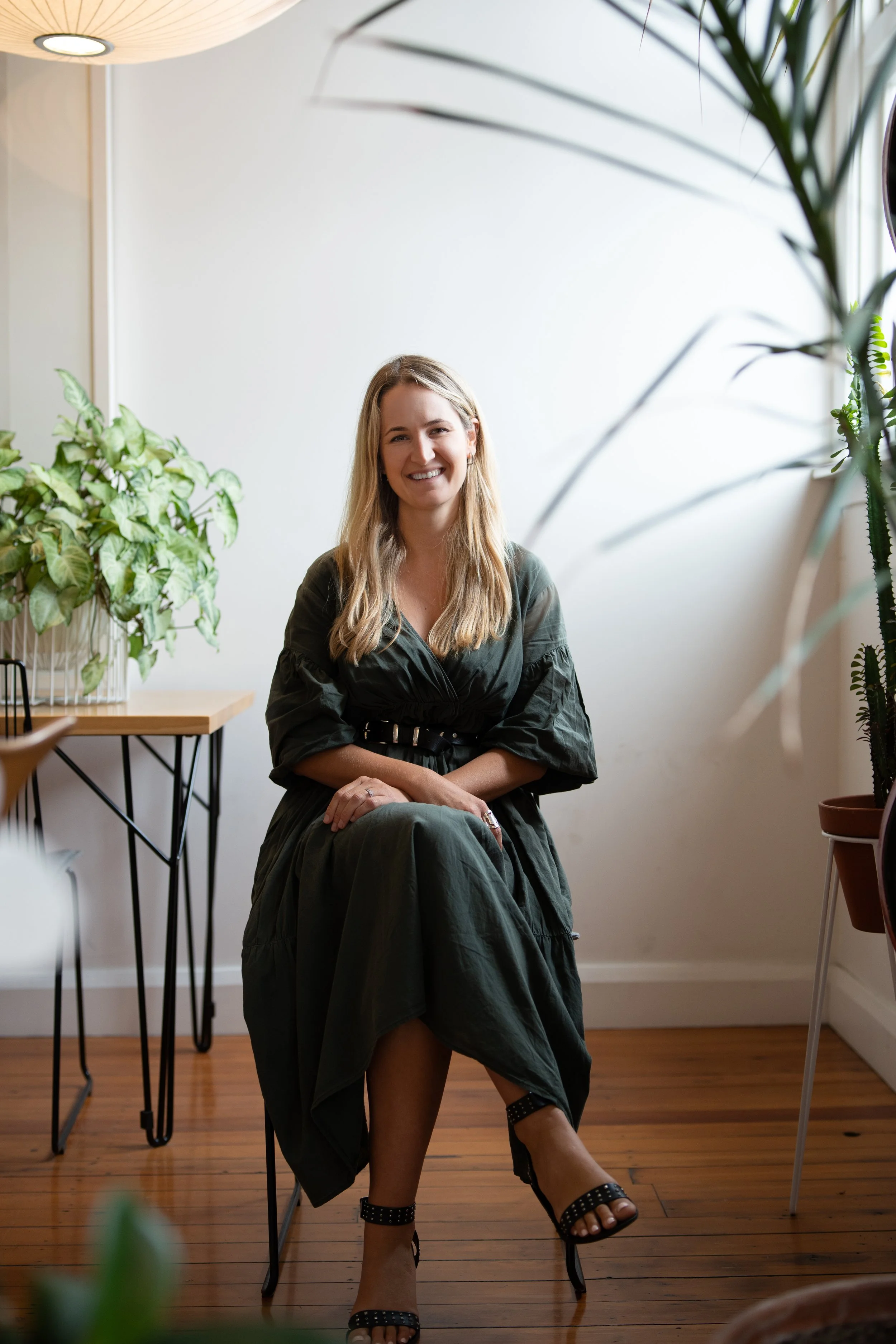 A woman sitting on a chair in a room with wooden floor, surrounded by potted plants, smiling at the camera.