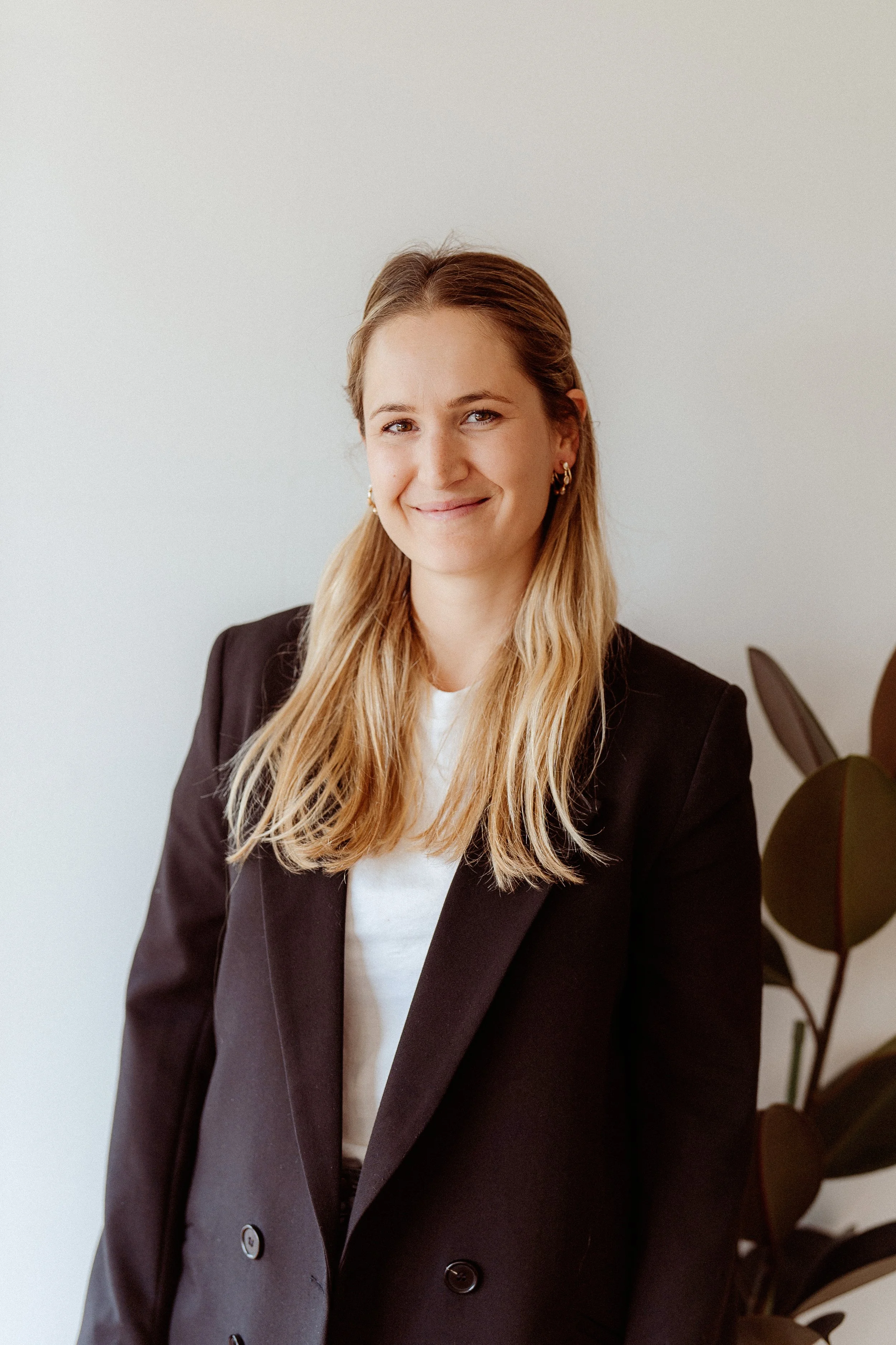 A woman with long blonde hair, wearing a black blazer, standing indoors next to a large plant, smiling at the camera.