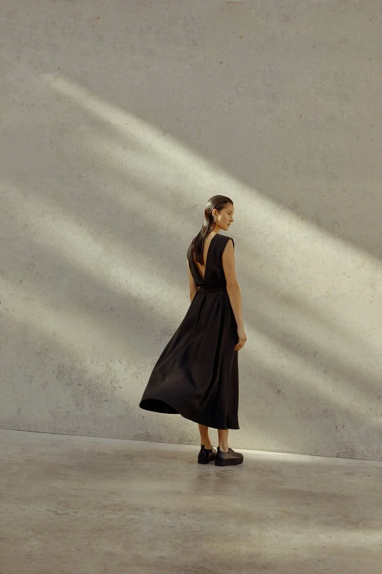 Woman in a long black dress standing against a textured gray wall with sunlight streaming in.