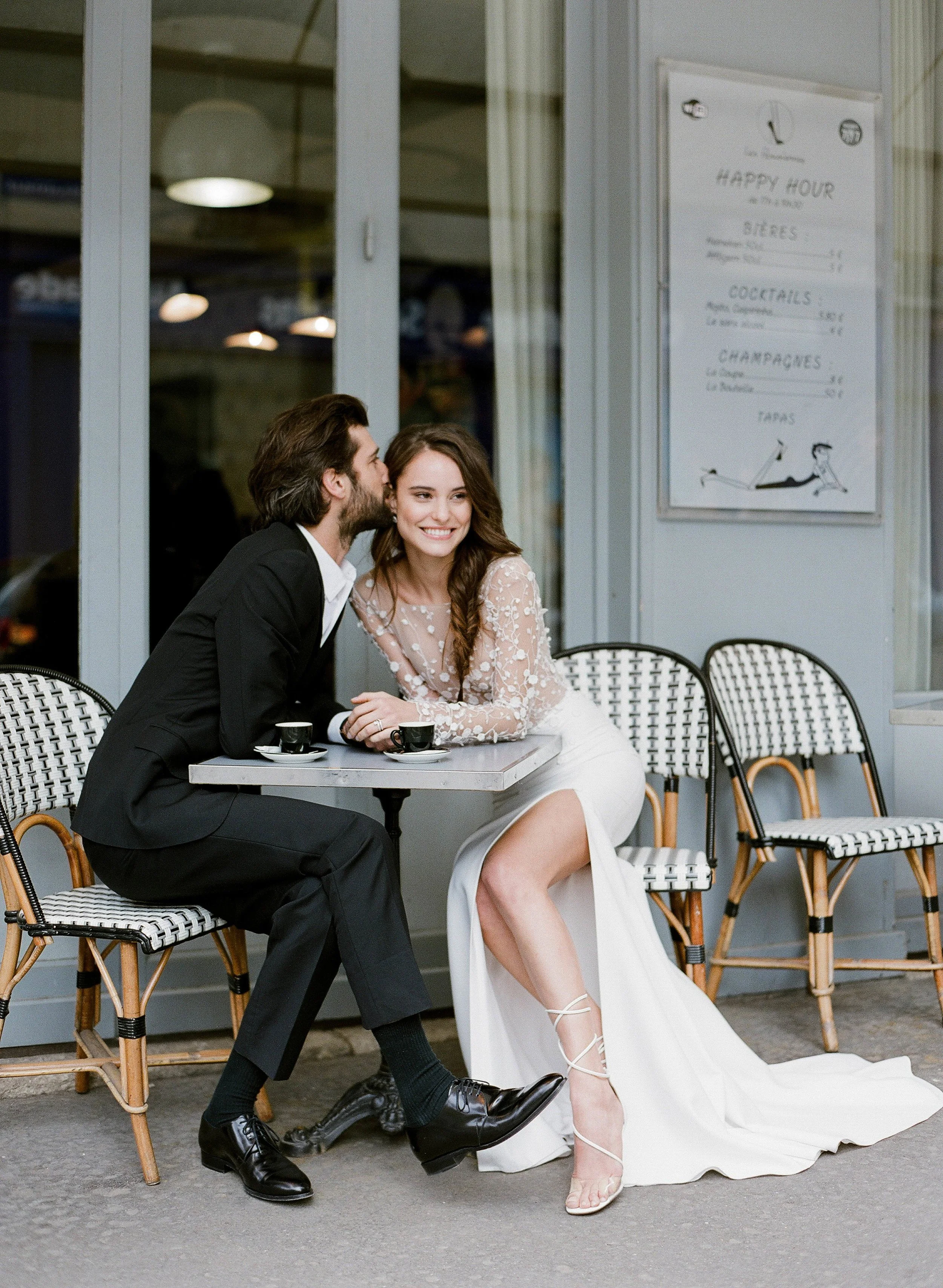 A happy couple sitting at an outdoor café table, with the man whispering into the woman's ear. The woman is smiling, wearing a white lace dress, and the man is wearing a black suit. There are coffee cups on the table, and a menu board in the background.