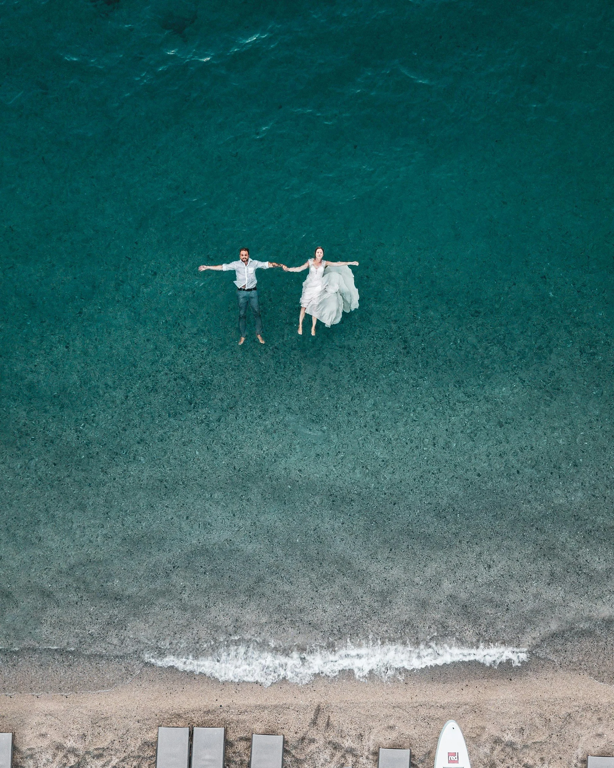 A couple in wedding attire lying on the beach holding hands, with the woman in a white wedding dress and the man in a white shirt and dark pants, seen from above.