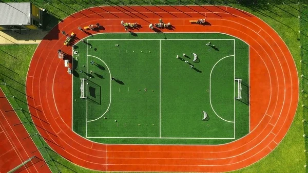 An aerial view of a sports field with a red running track surrounding a green soccer field, with people practicing soccer.