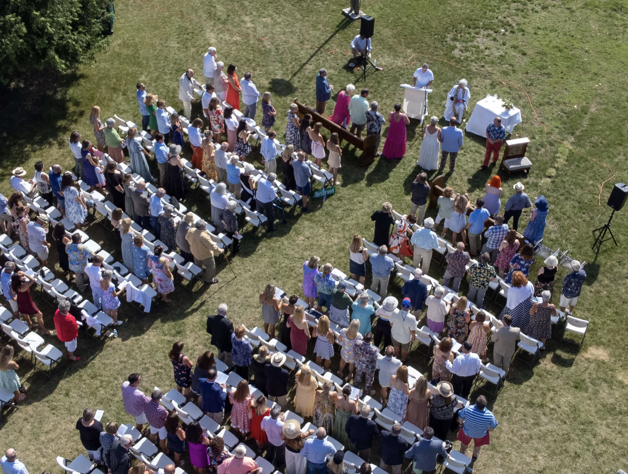 An aerial view of a large outdoor wedding ceremony with people standing and sitting on white chairs on green grass. Guests are dressed in bright summer clothing, and to the front, a few people are gathered near an altar with a table and flowers.