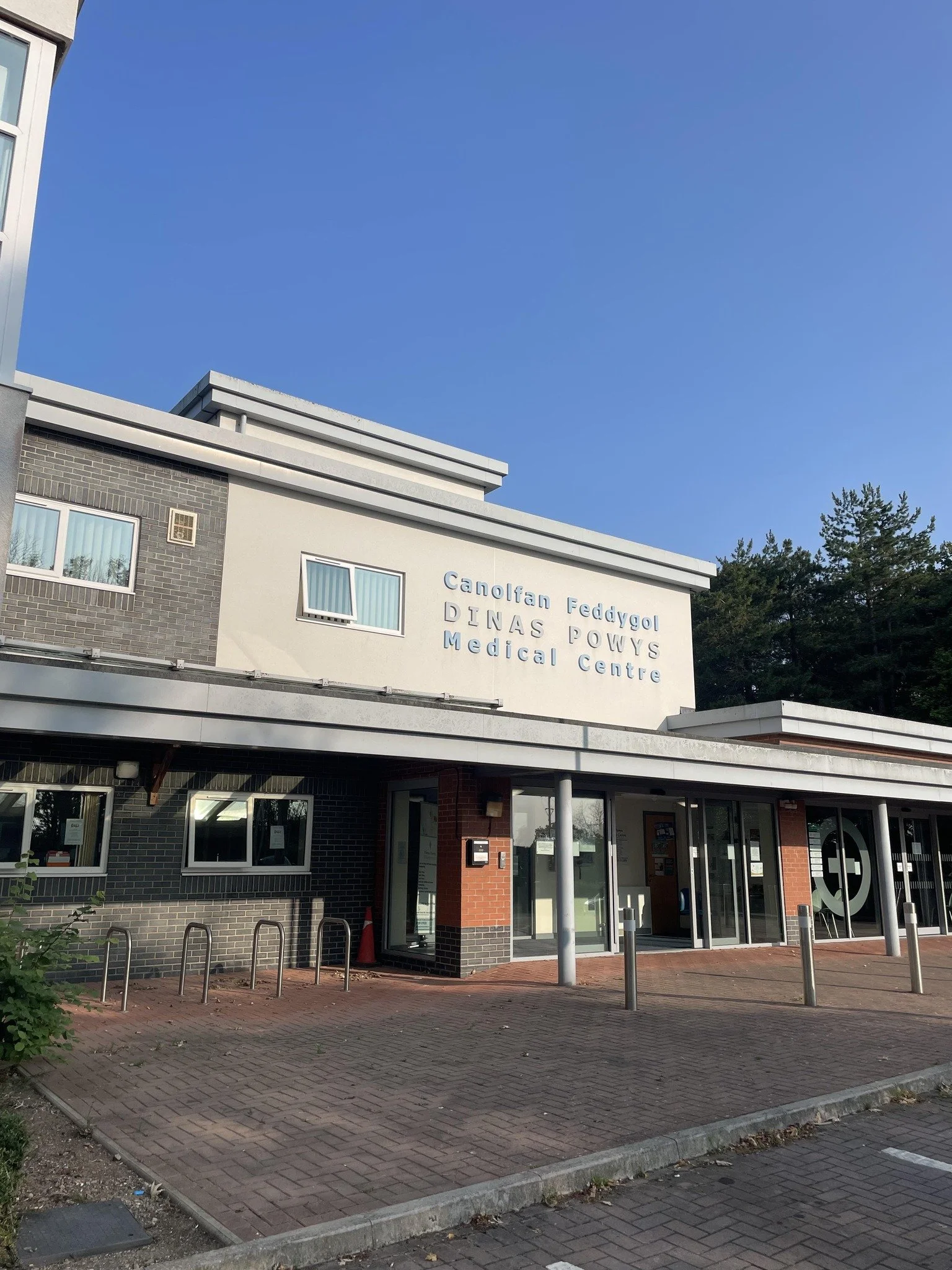 Exterior view of Dinas Powys medical center building with a sign that reads 'Canolfan Fedygol DINAS POWYS Medical Centre,' featuring brick and light-colored walls, windows, a glass entrance, and bicycle racks outside under a clear blue sky.