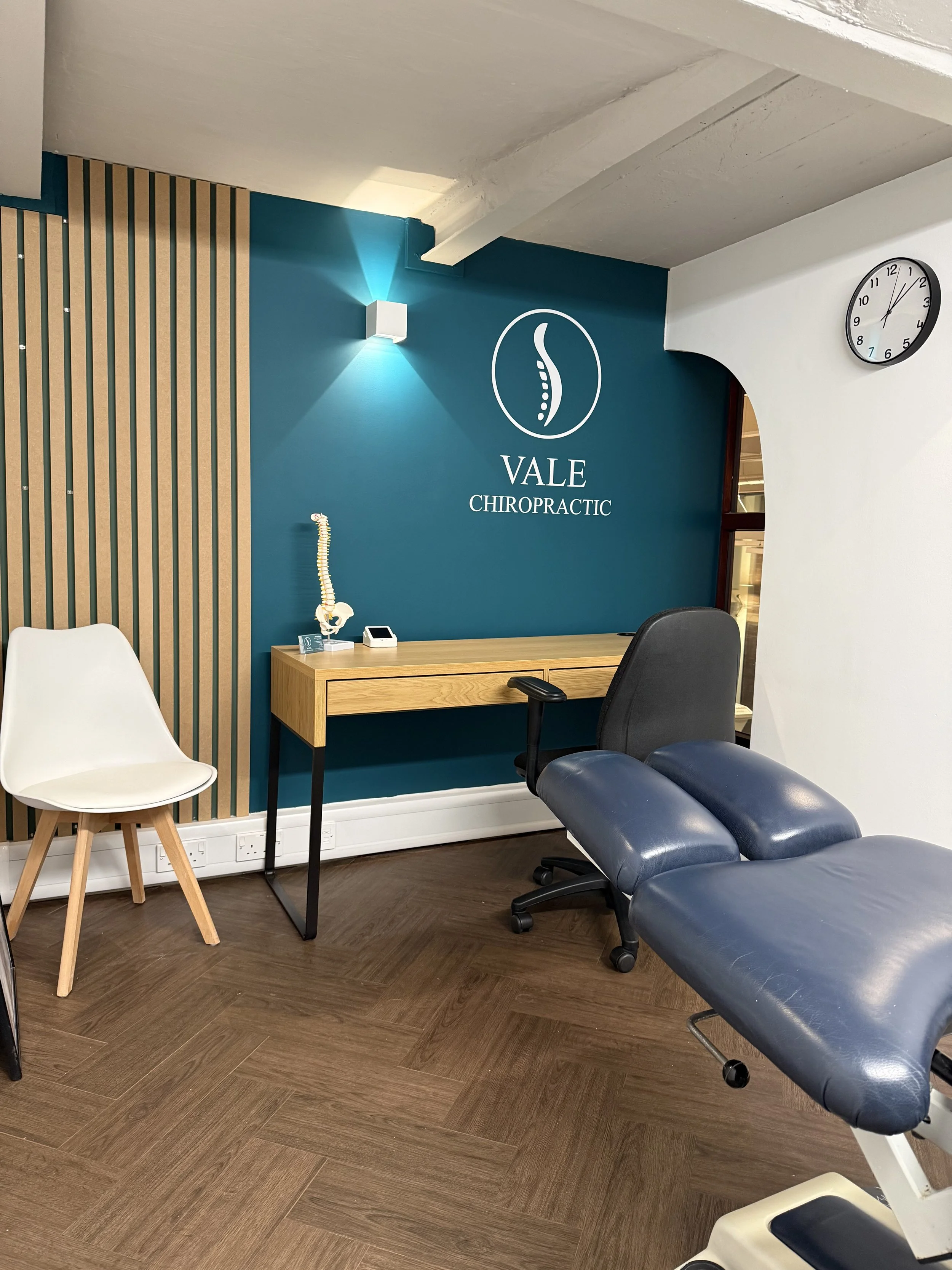 Interior of VALE Chiropractic clinic with a treatment table, chair, a wooden desk with a model of a human spine, and a wall clock showing 1:07.