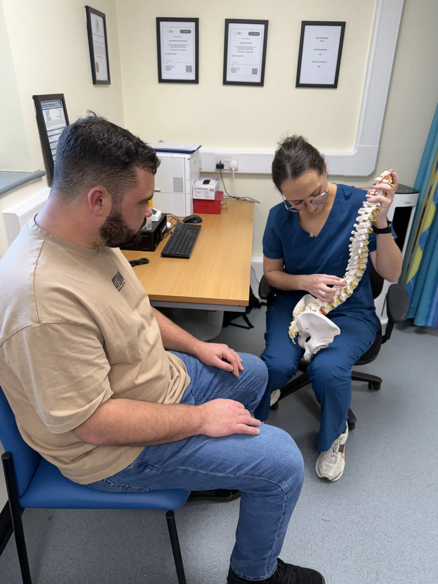 A healthcare professional in blue scrubs showing a model of a human spine to a man sitting in a chair in a medical office.