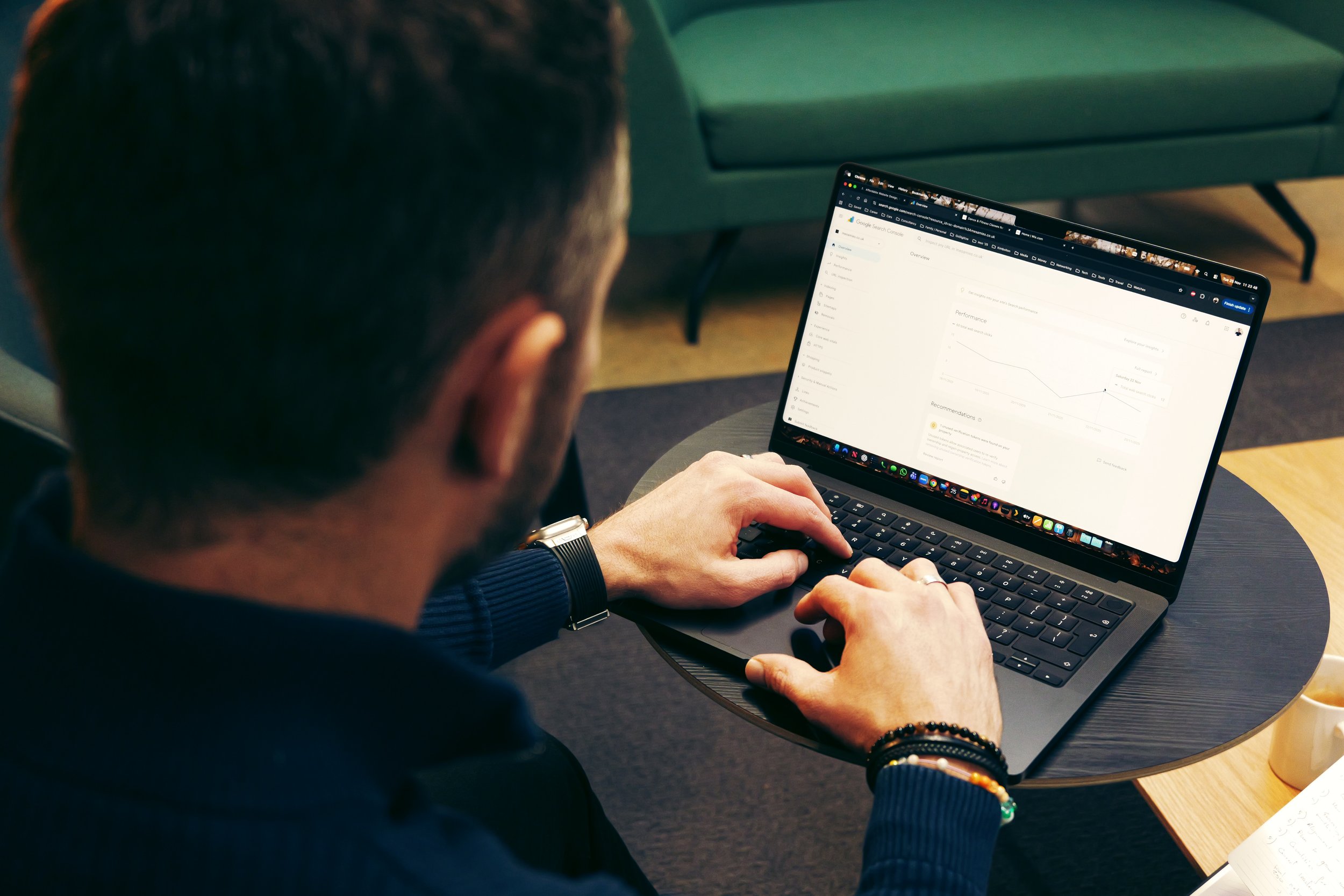 Alex working on a laptop at a round table, with a green sofa in the background. The screen displays a performance analytics chart and various web browser tabs.