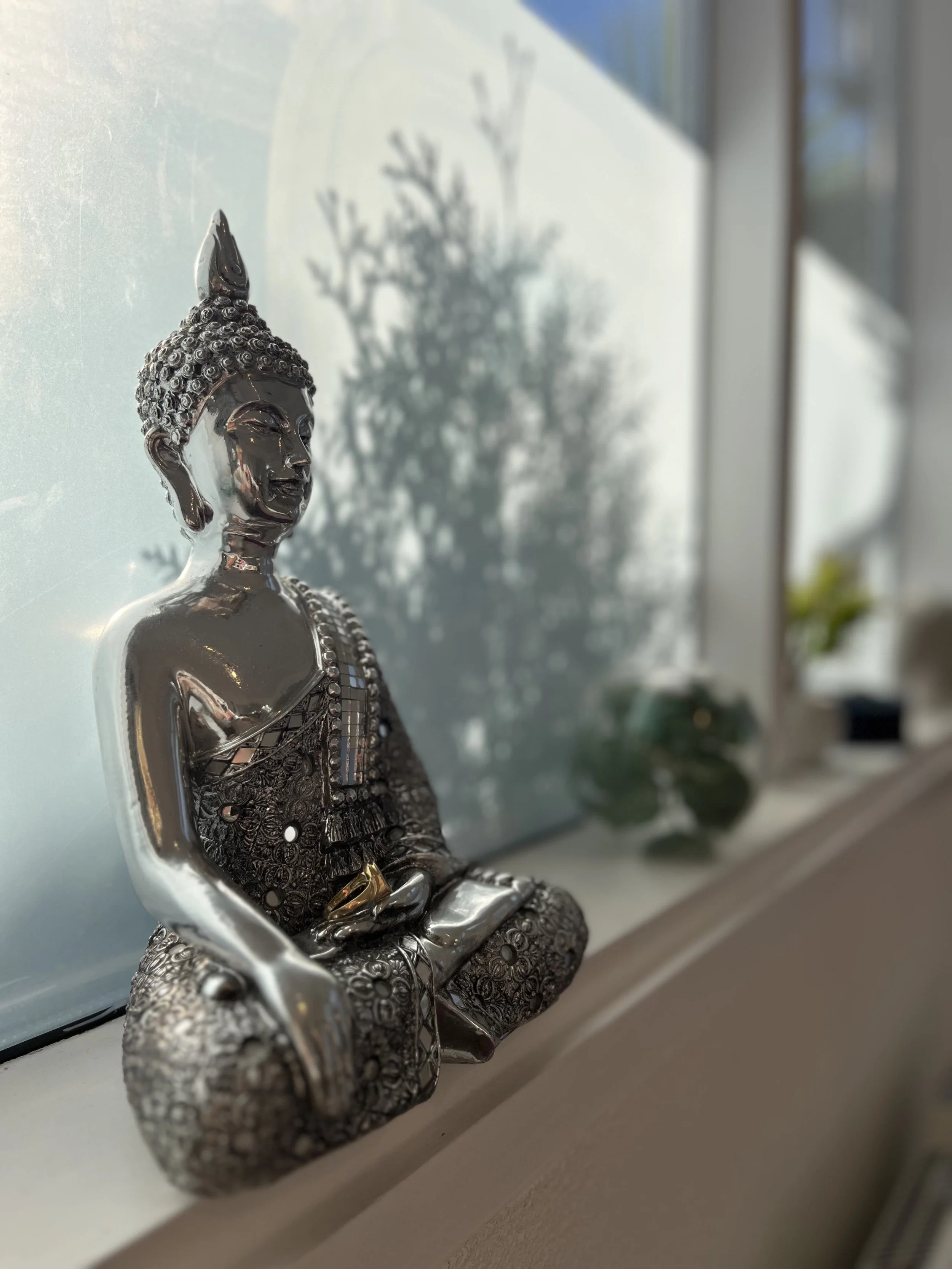 A shiny metallic Buddha statue sitting on a windowsill, casting a shadow of a tree on the window.