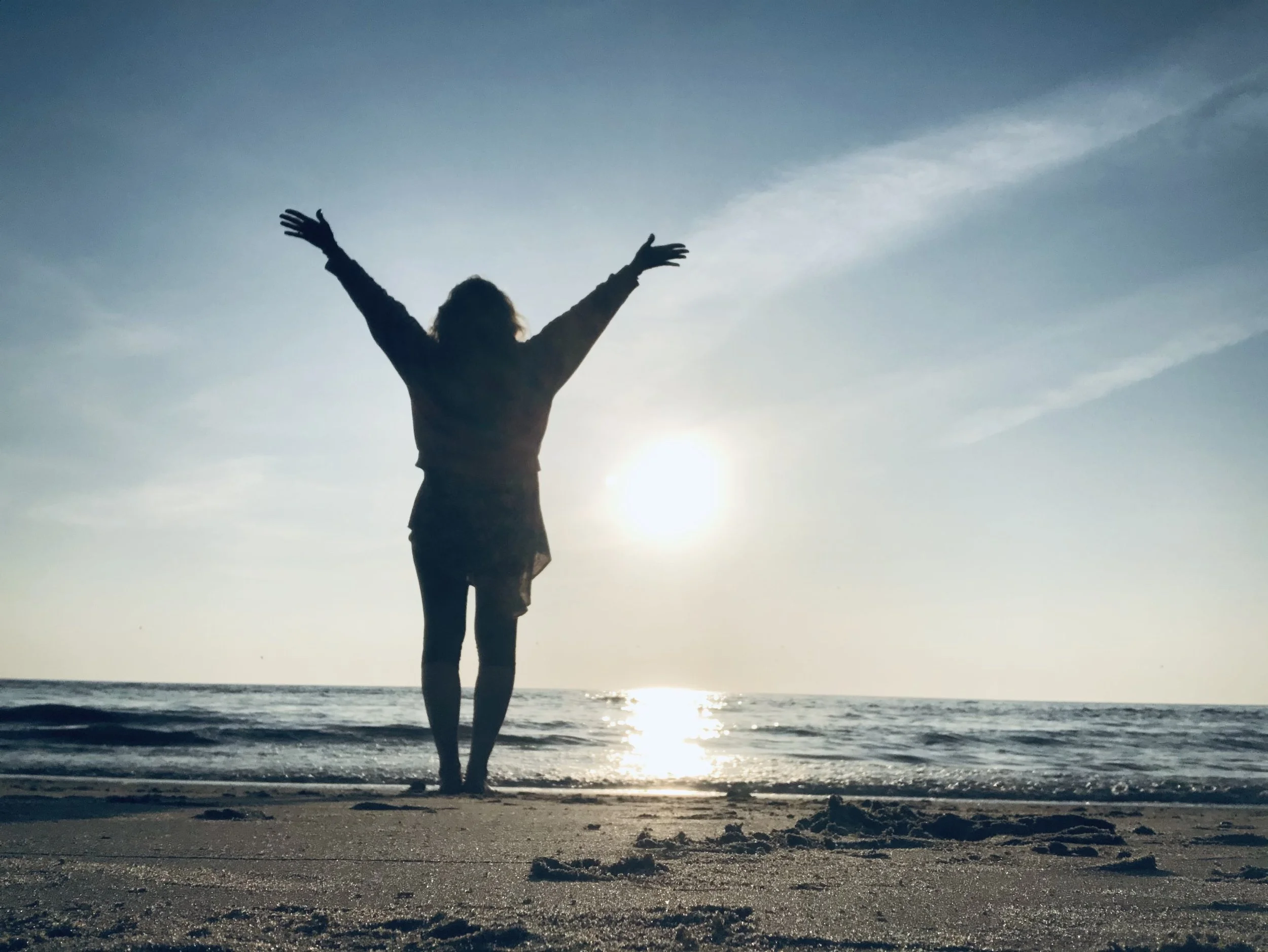 Silhouette of a woman standing on the beach with her arms raised, facing the sunset over the ocean.
