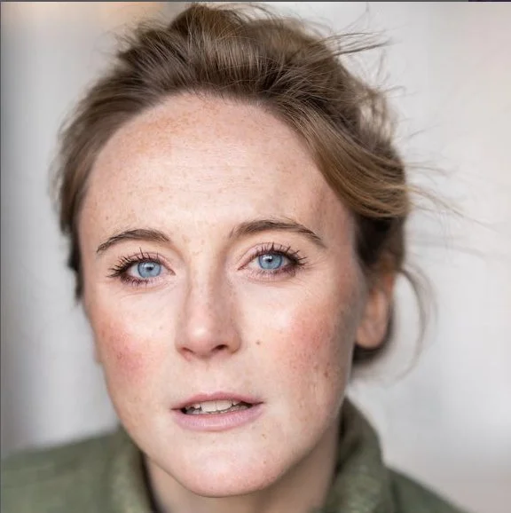 Close-up portrait of a woman with blue eyes and light skin, looking directly at the camera against a plain background.