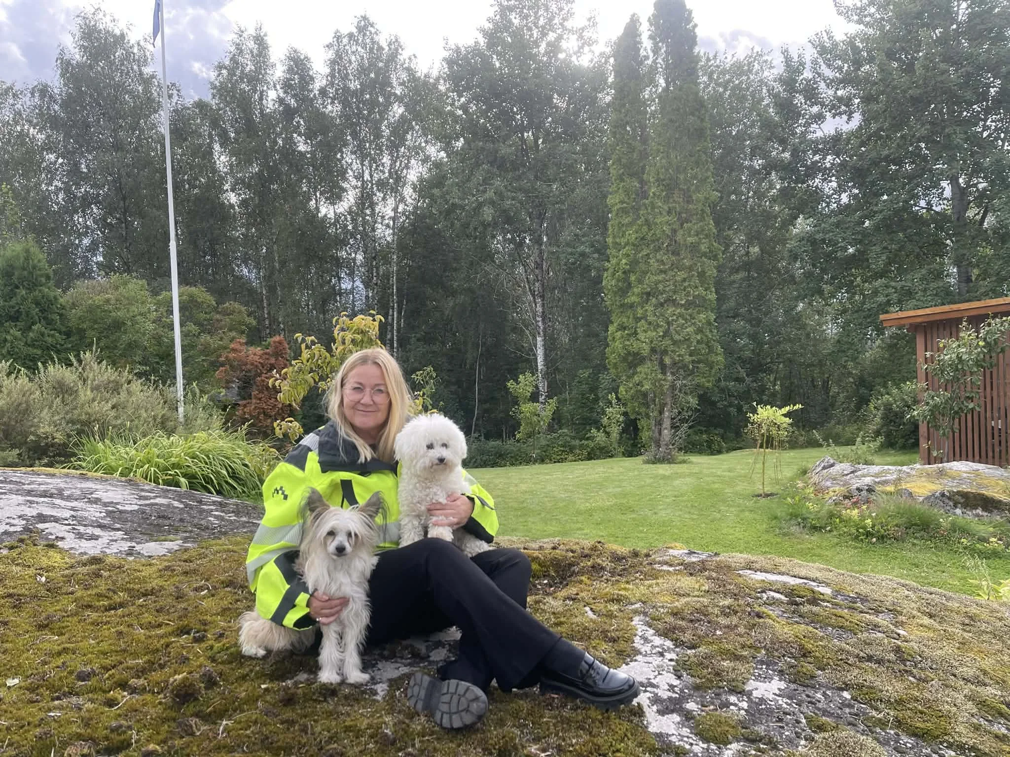 En kvinne i en gul og svart jakke sitter på en stein i en grønn hage, holder to hunder. En liten, langhåret hund hvit og grå, og en større, hvit, fluffy hund.