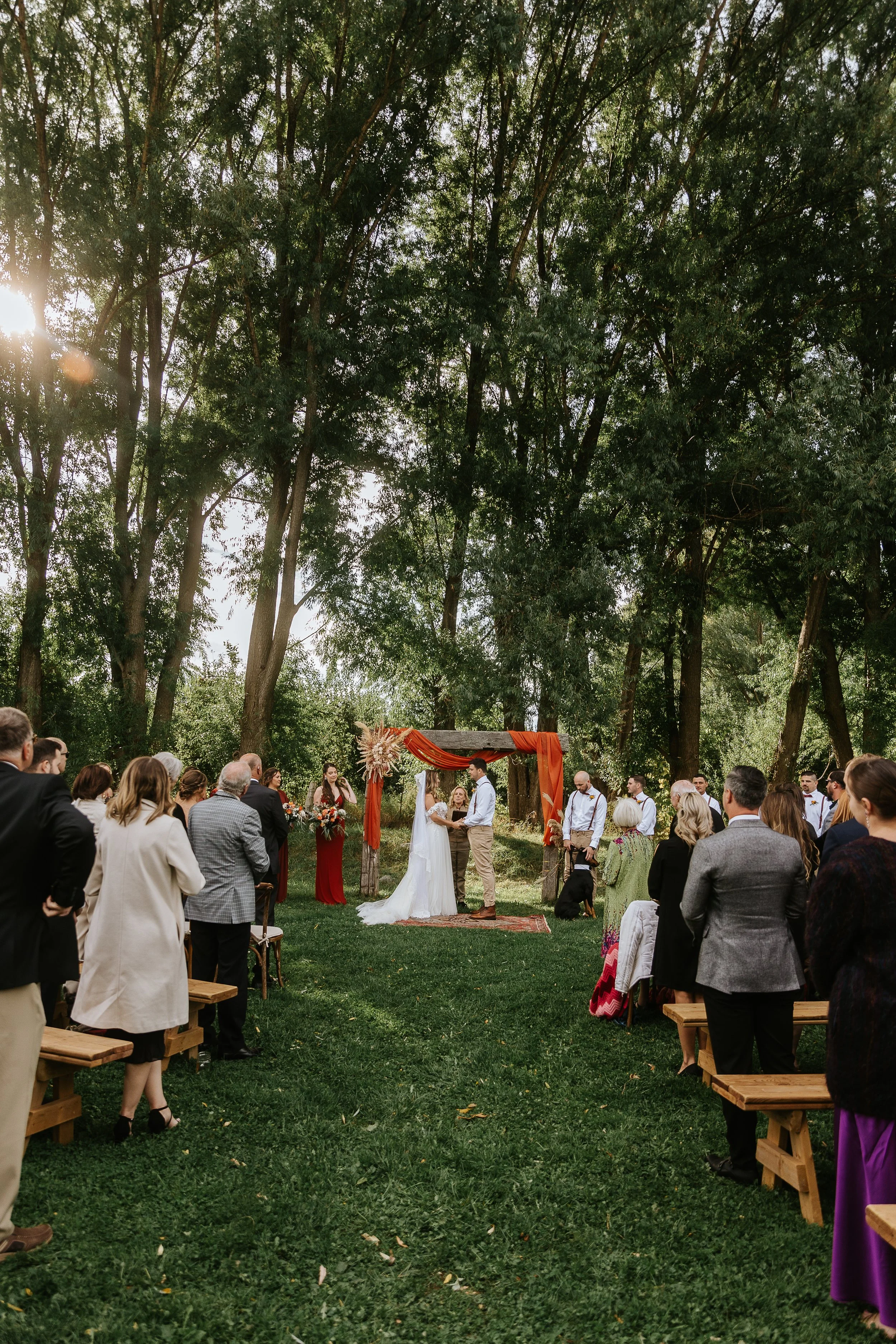 Bride and groom exchanging vows at an outdoor wedding in Hamilton Ontario, surrounded by guests and lush trees – James and Alyson Photography