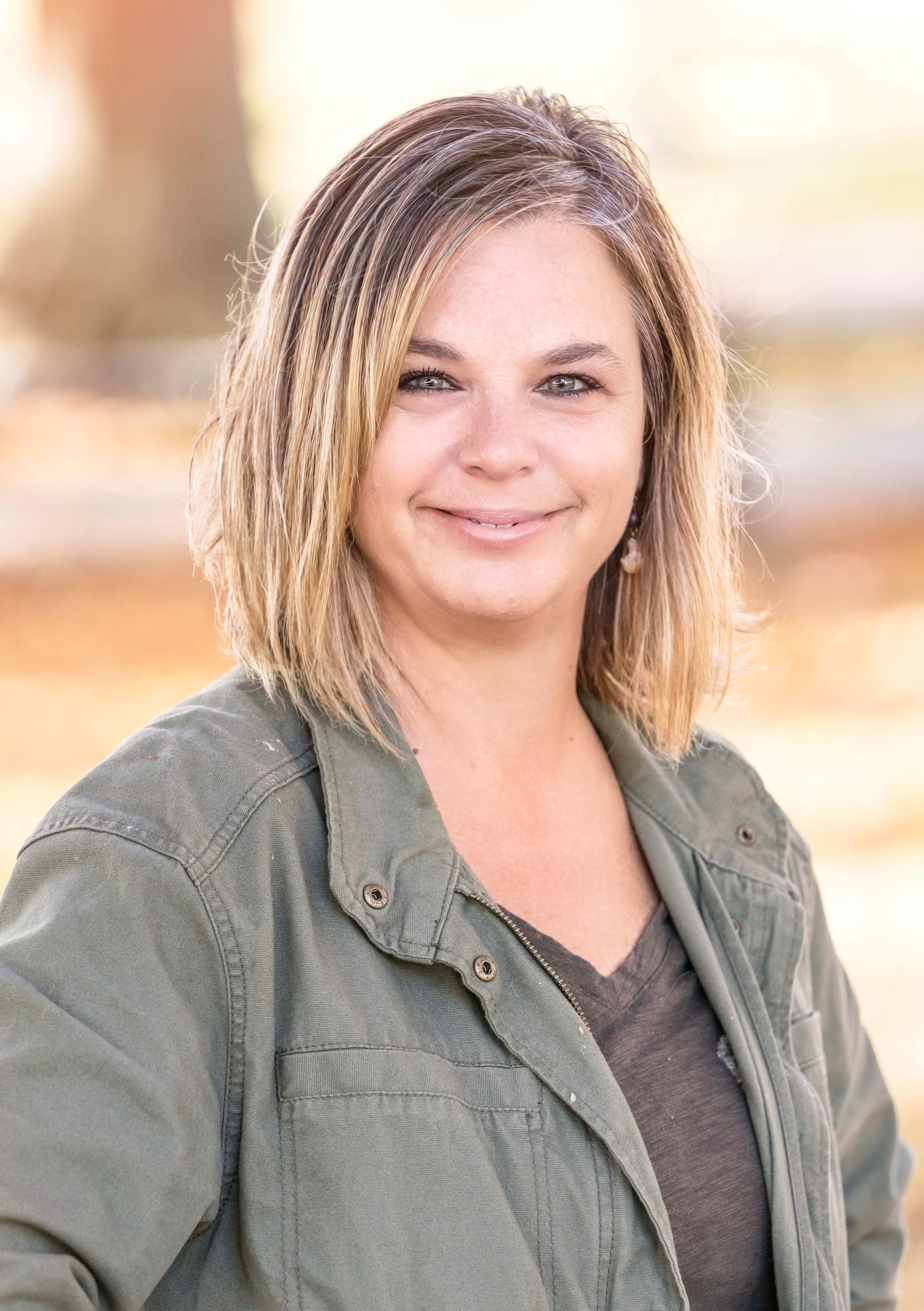 A woman with shoulder-length light brown hair and grey eyes, smiling outdoors during fall, wearing a green jacket and earrings.