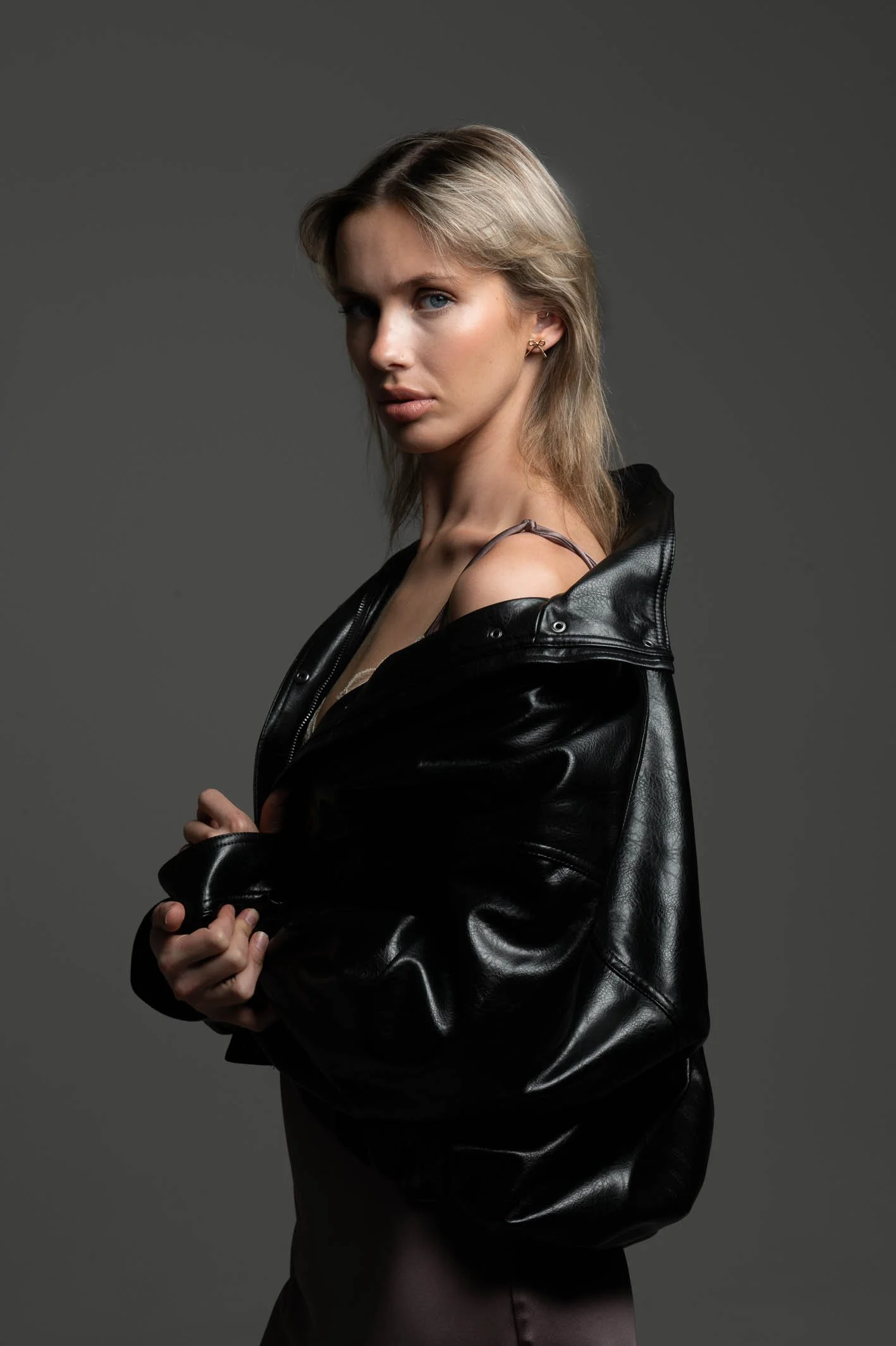 A woman with blond hair and blue eyes wearing a black leather jacket and a dark dress, standing against a plain gray background.