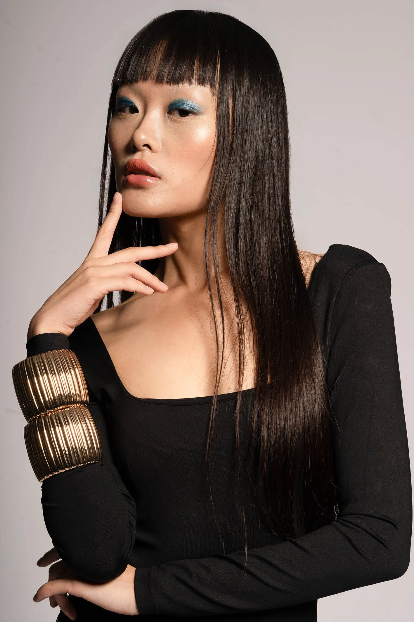 A woman with long dark hair and bangs, wearing a black top and gold jewelry, posing with her hand near her face against a light background.