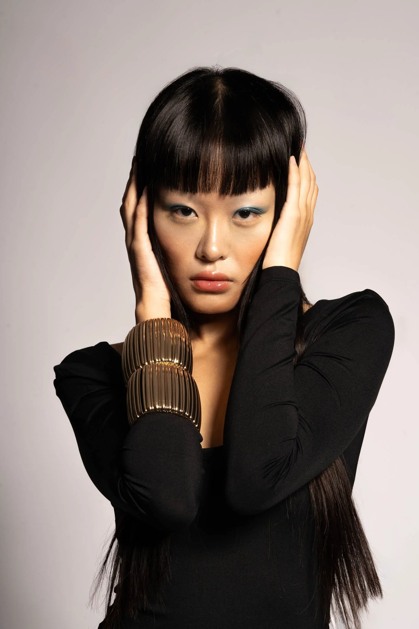A woman with straight black hair and bangs, wearing a black top and large gold bracelets, posing with her hands on her head against a plain light background.