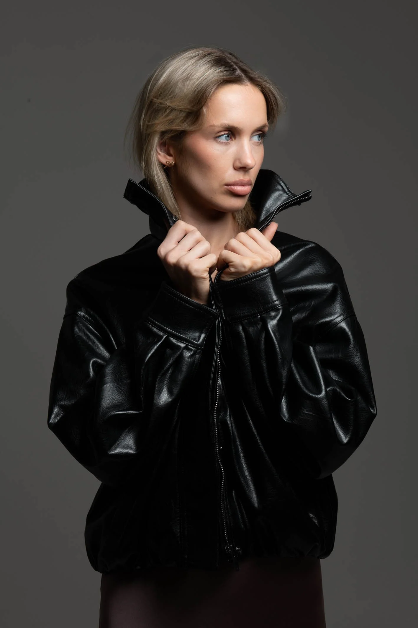 A young woman with blonde hair and blue eyes wearing a black leather jacket, holding the collar with both hands, looking to her left against a plain grey background.