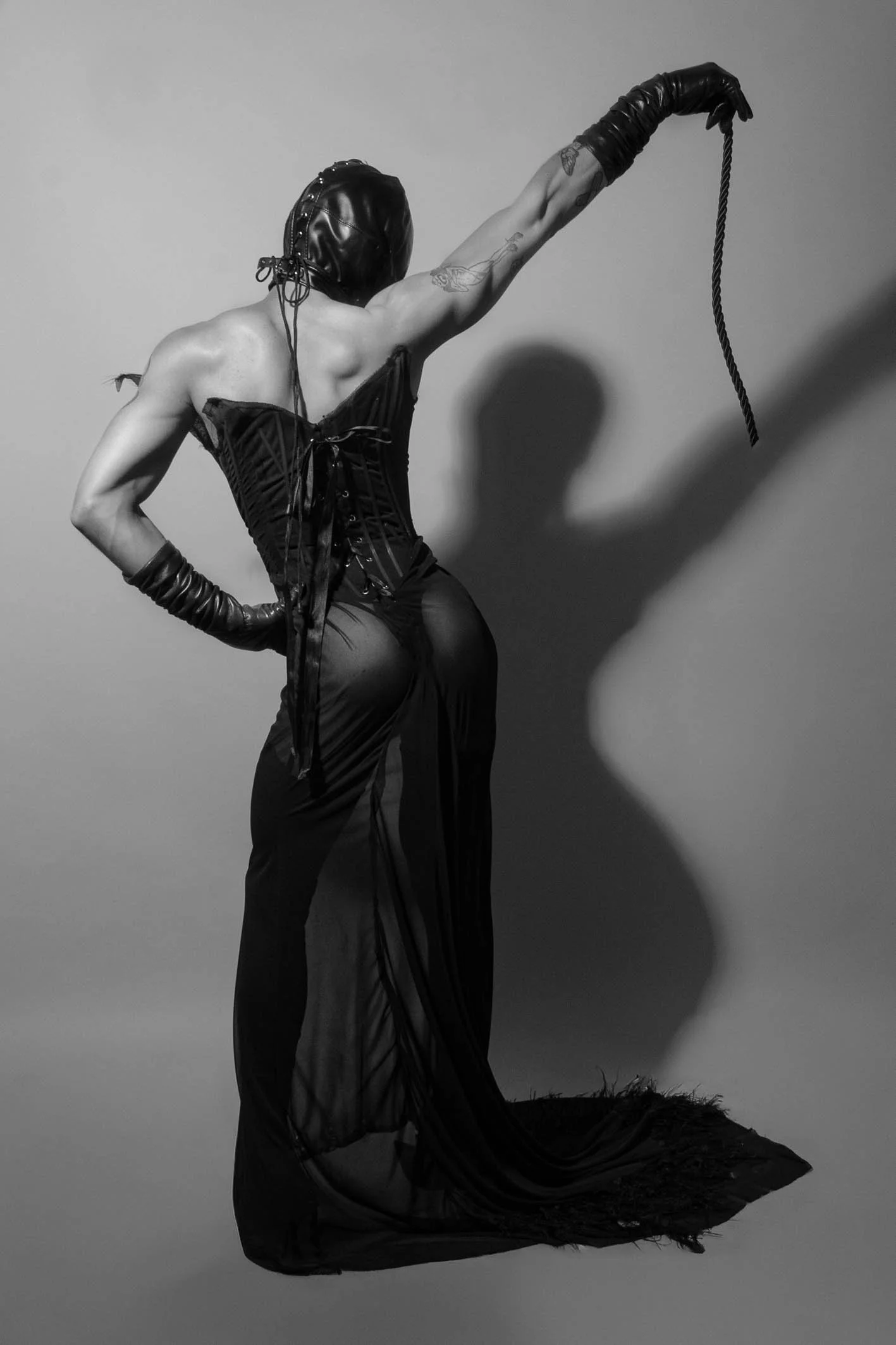 A person in gothic attire with a hood, corset, long gloves, and sheer skirt, holding a whip, standing against a plain background with a shadow cast on the wall.