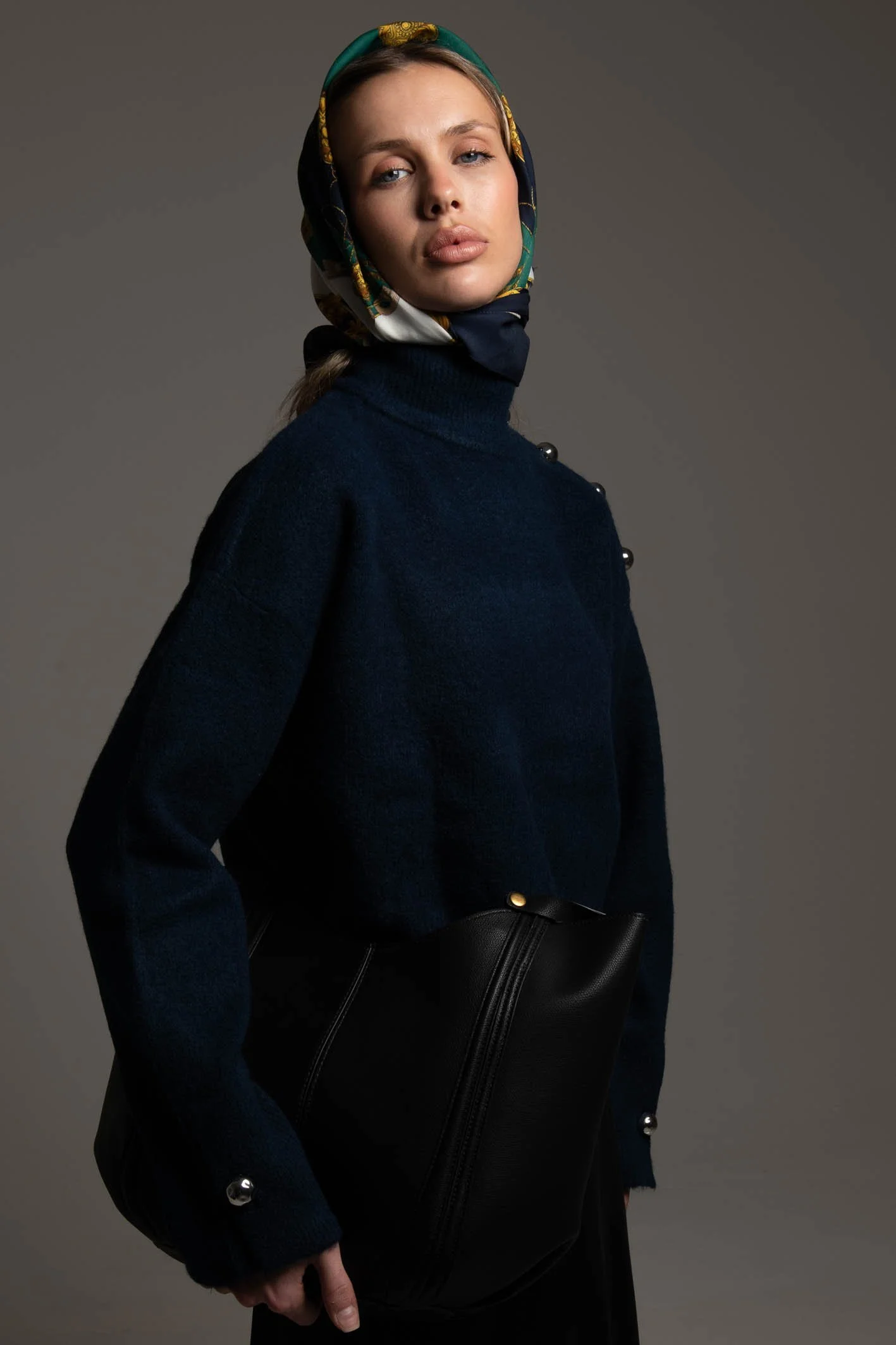 A woman wearing a dark blue turtleneck sweater with pearl button accents, a patterned silk headscarf, and carrying a black leather bag against a neutral background.