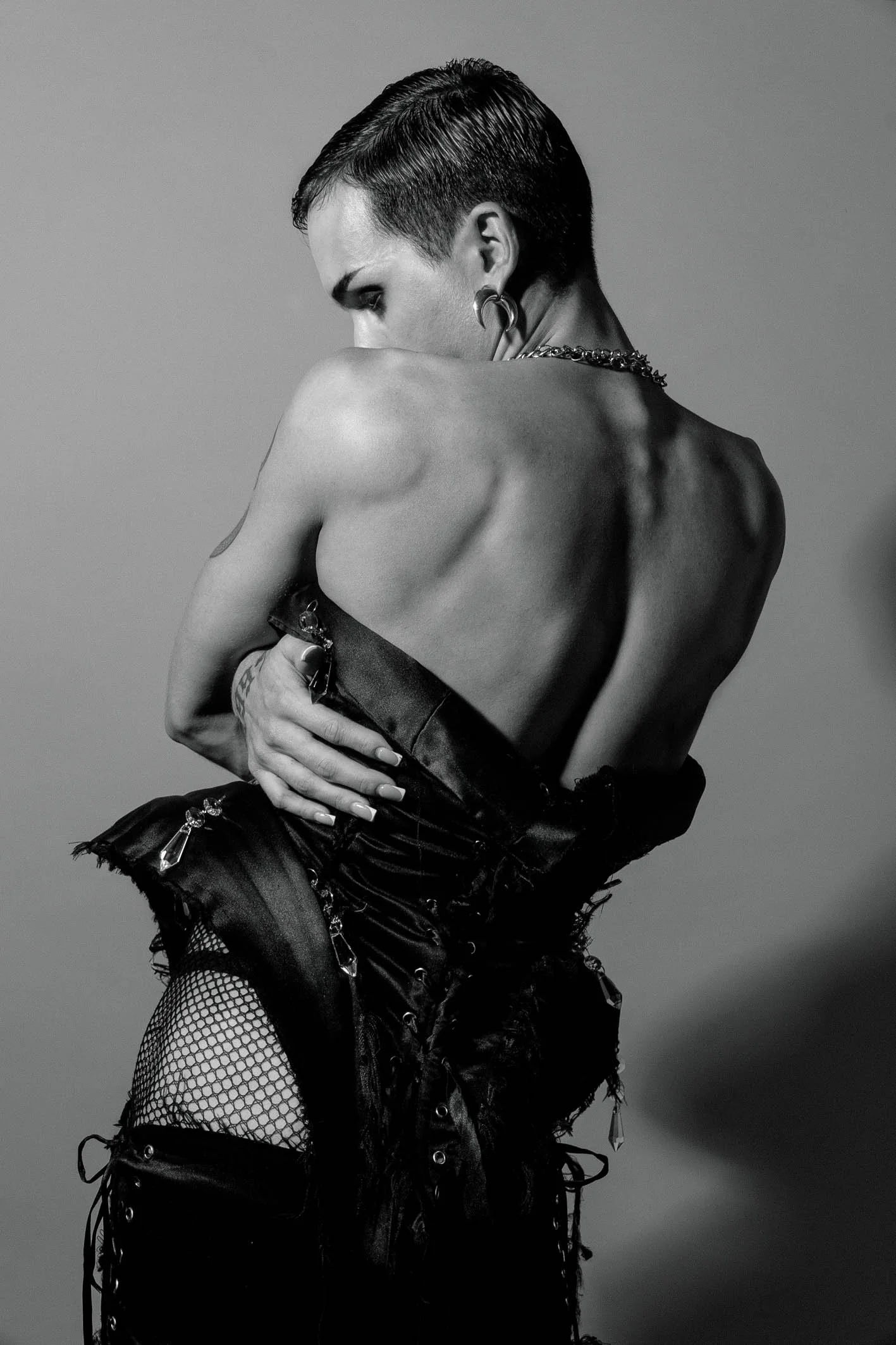 A person with short hair and multiple earrings, wearing a distressed black leather outfit with fishnet stockings, posing with her back partially turned and her face partially obscured in a black and white photograph.