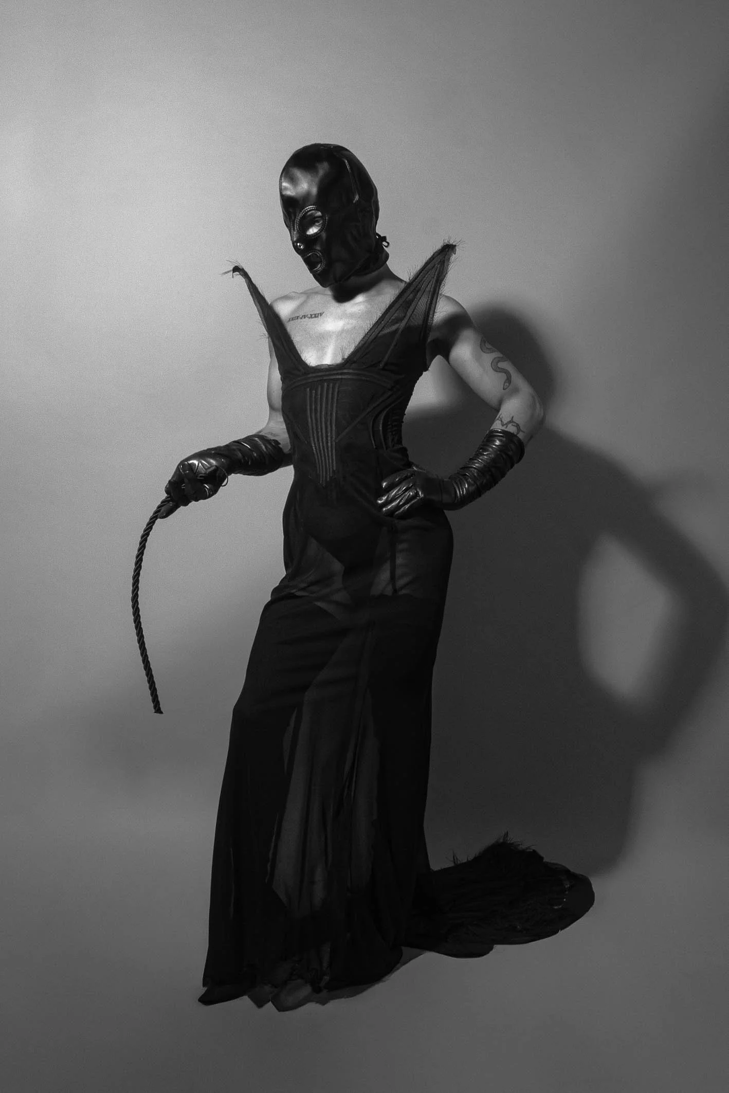 A person dressed in a black, gothic outfit with a full-face black mask, gloves, and long dress, holding a whip, standing against a plain background.