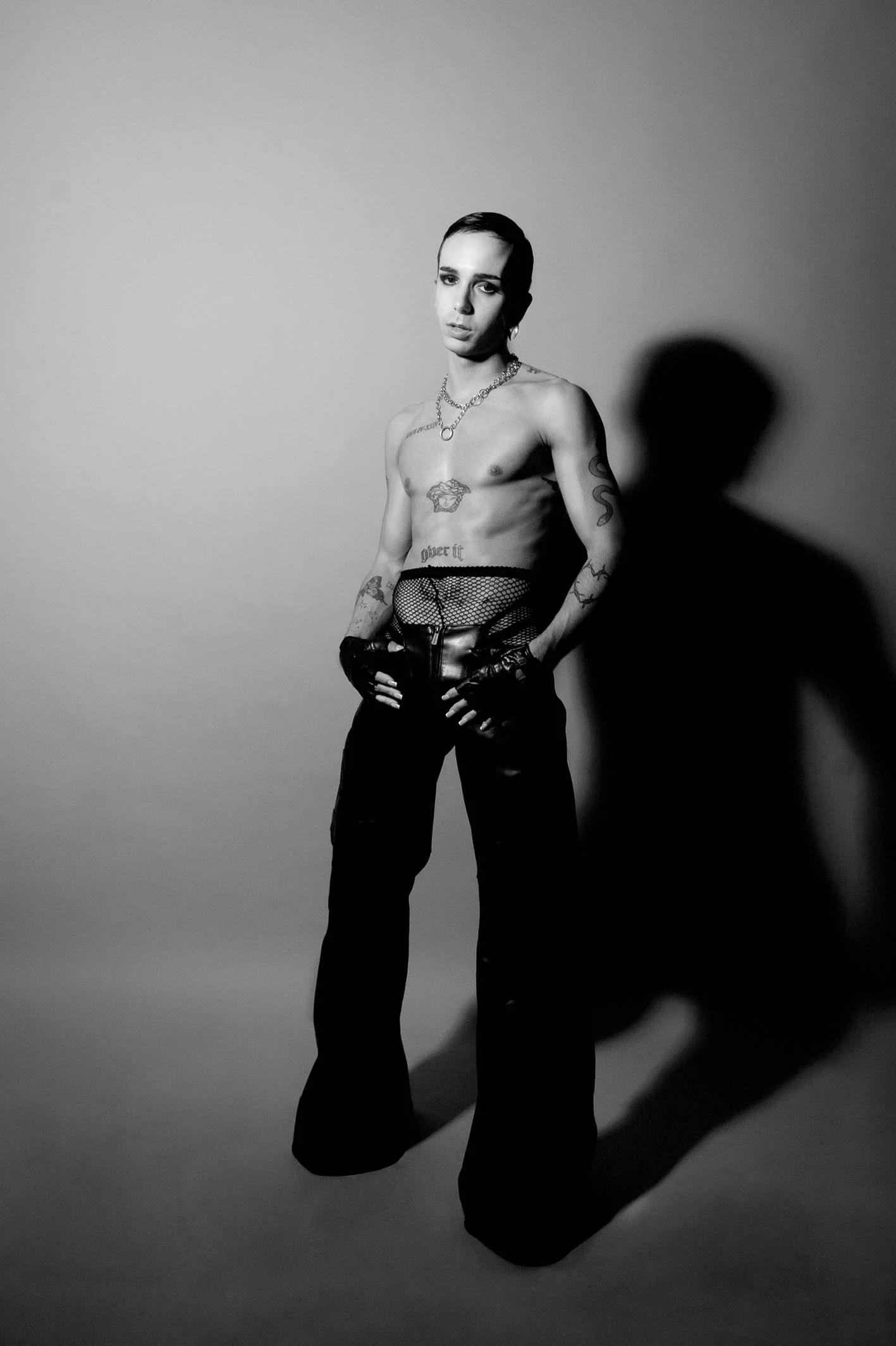 A black-and-white photo of a shirtless person with tattoos, wearing fishnet gloves and high-waisted pants, standing against a plain background with a shadow cast to their right.