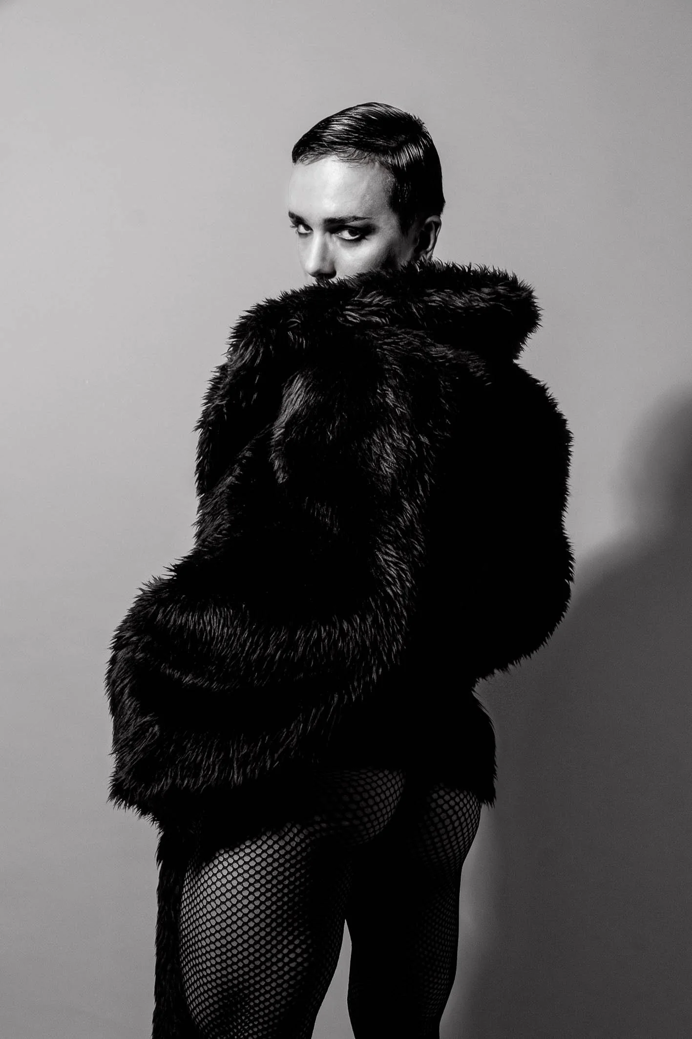A person in a shiny black fur coat and fishnet stockings looking over her shoulder, with her face partially obscured by her coat.
