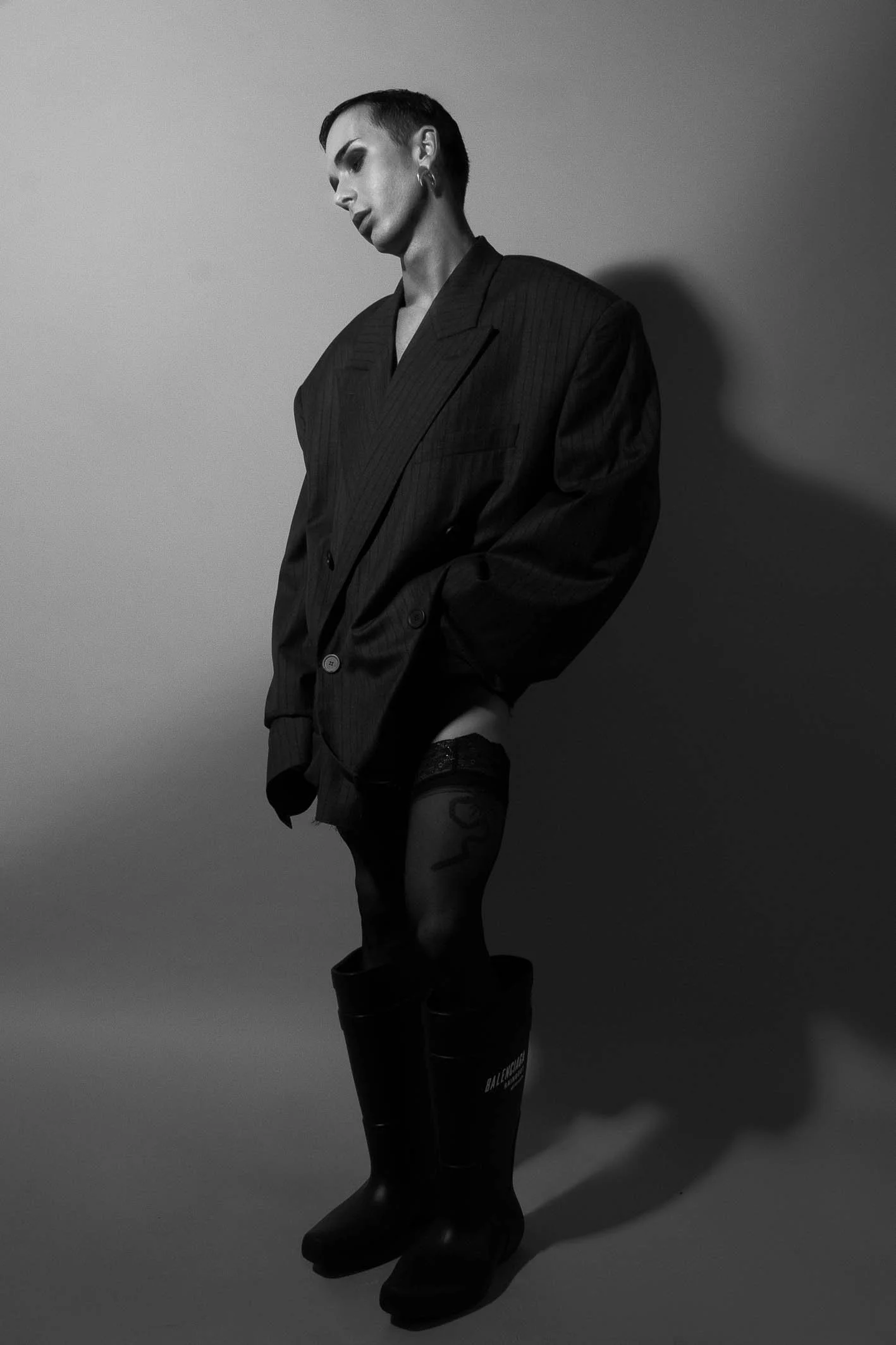 Black and white photo of a person with short hair wearing an oversized blazer, thigh-high socks, and rain boots, posing against a plain wall.