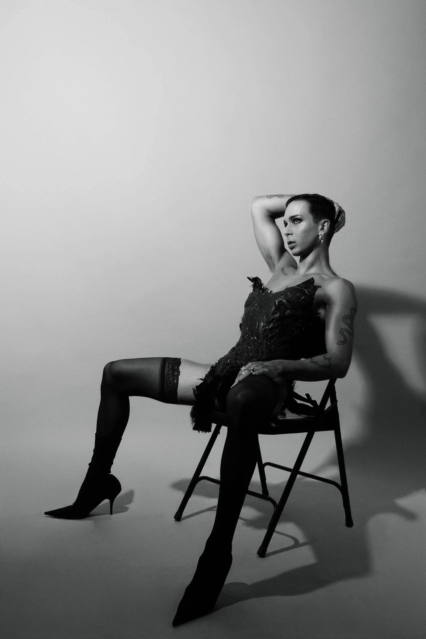 A black-and-white photo of a woman sitting on a chair, wearing a dress, thigh-high stockings, and high heels. She has short hair, tattoos on her arms, and is posing with one arm behind her head, looking to the right.