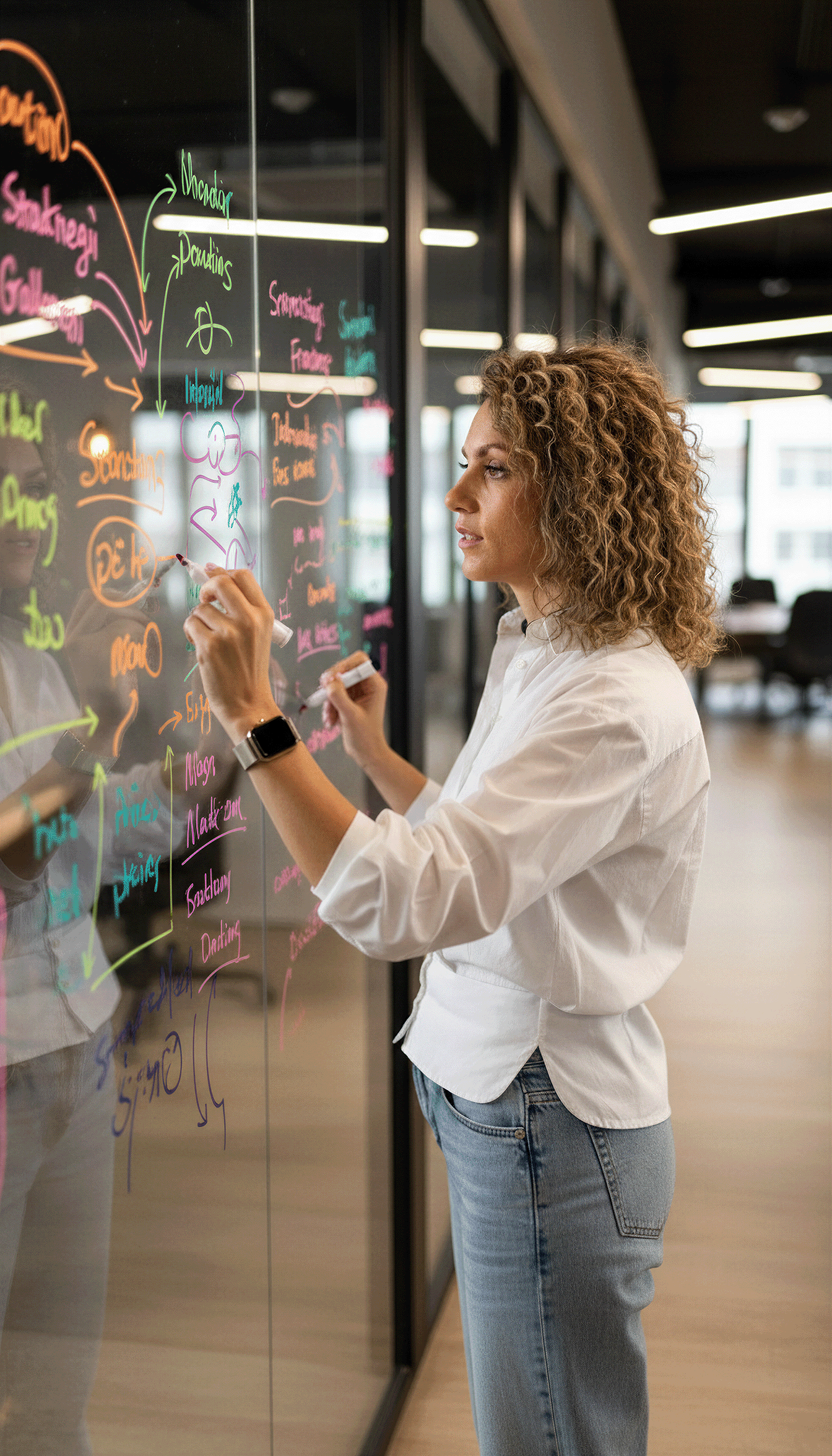 A woman with curly hair writing and drawing on a glass wall with colorful markers in an office